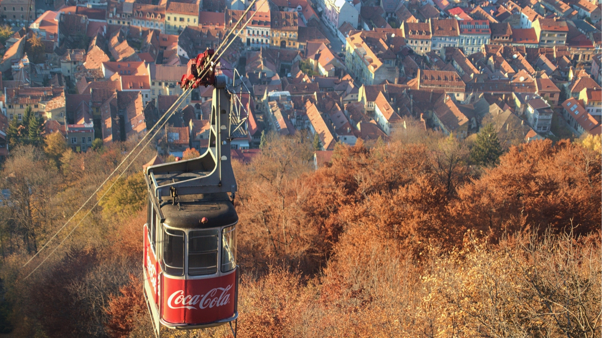 Aerial cable car with a Coca-Cola ad descends over autumn trees towards a sunlit town with red-tiled roofs, creating a warm, nostalgic atmosphere.