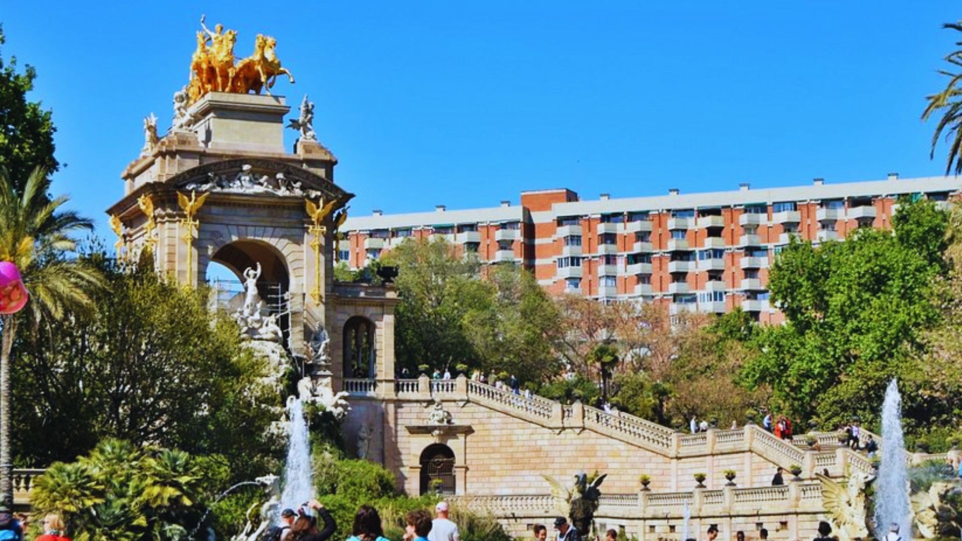 A vibrant scene at Barcelona's Parc de la Ciutadella features the ornate, golden Cascade Fountain under a clear blue sky, with visitors enjoying the sunny day.