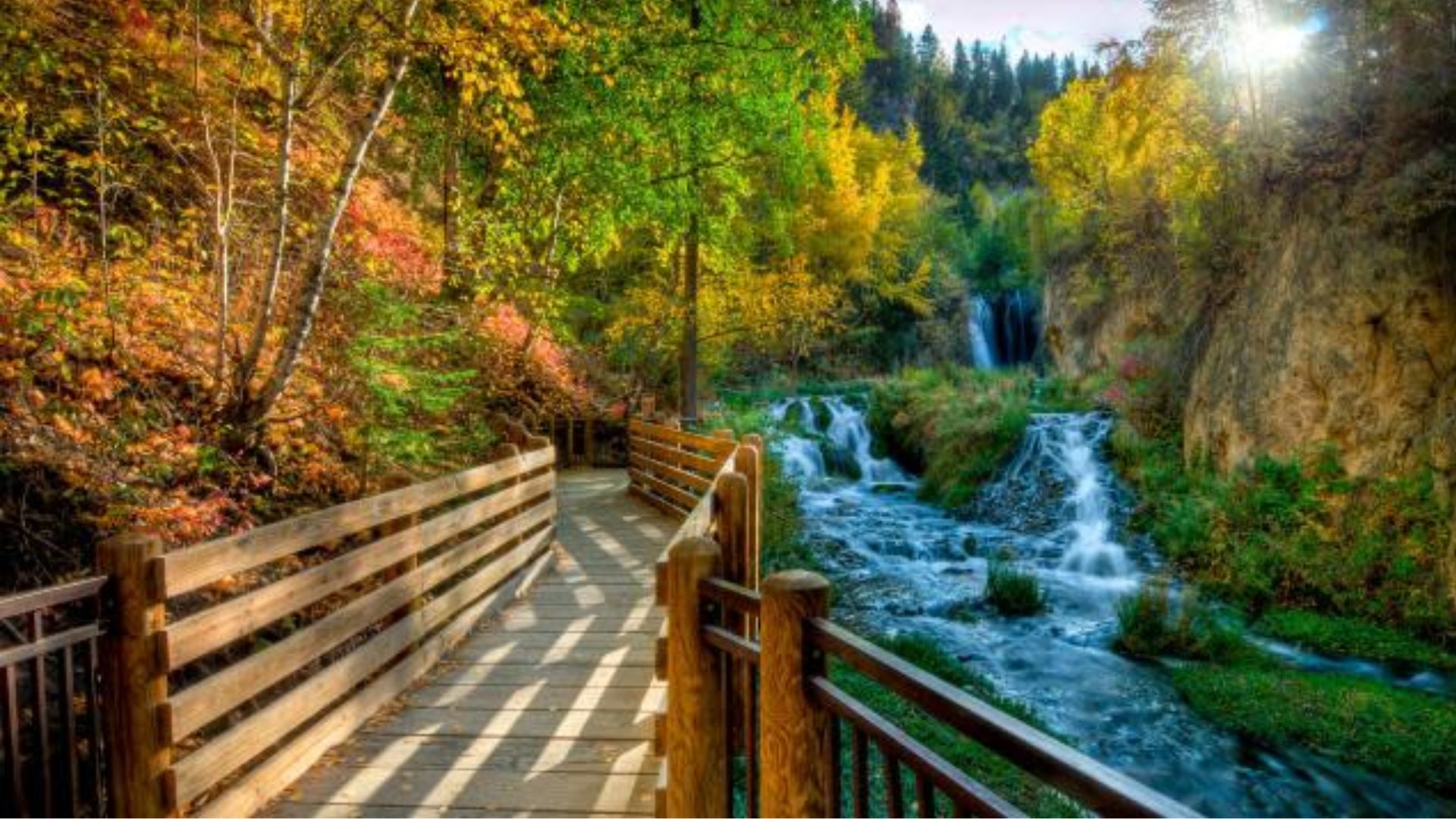 A wooden path curves through a vibrant autumn landscape with colorful trees. A cascading waterfall flows beside the path, creating a serene atmosphere.