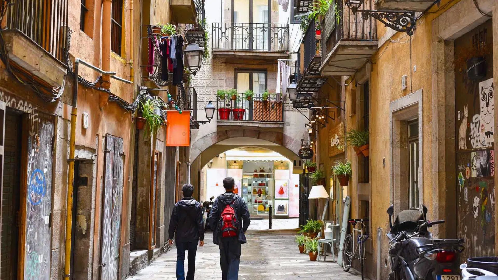 A narrow, picturesque alley in a European city features colorful buildings, balconies with plants, and two people walking away. The scene feels cozy and vibrant.