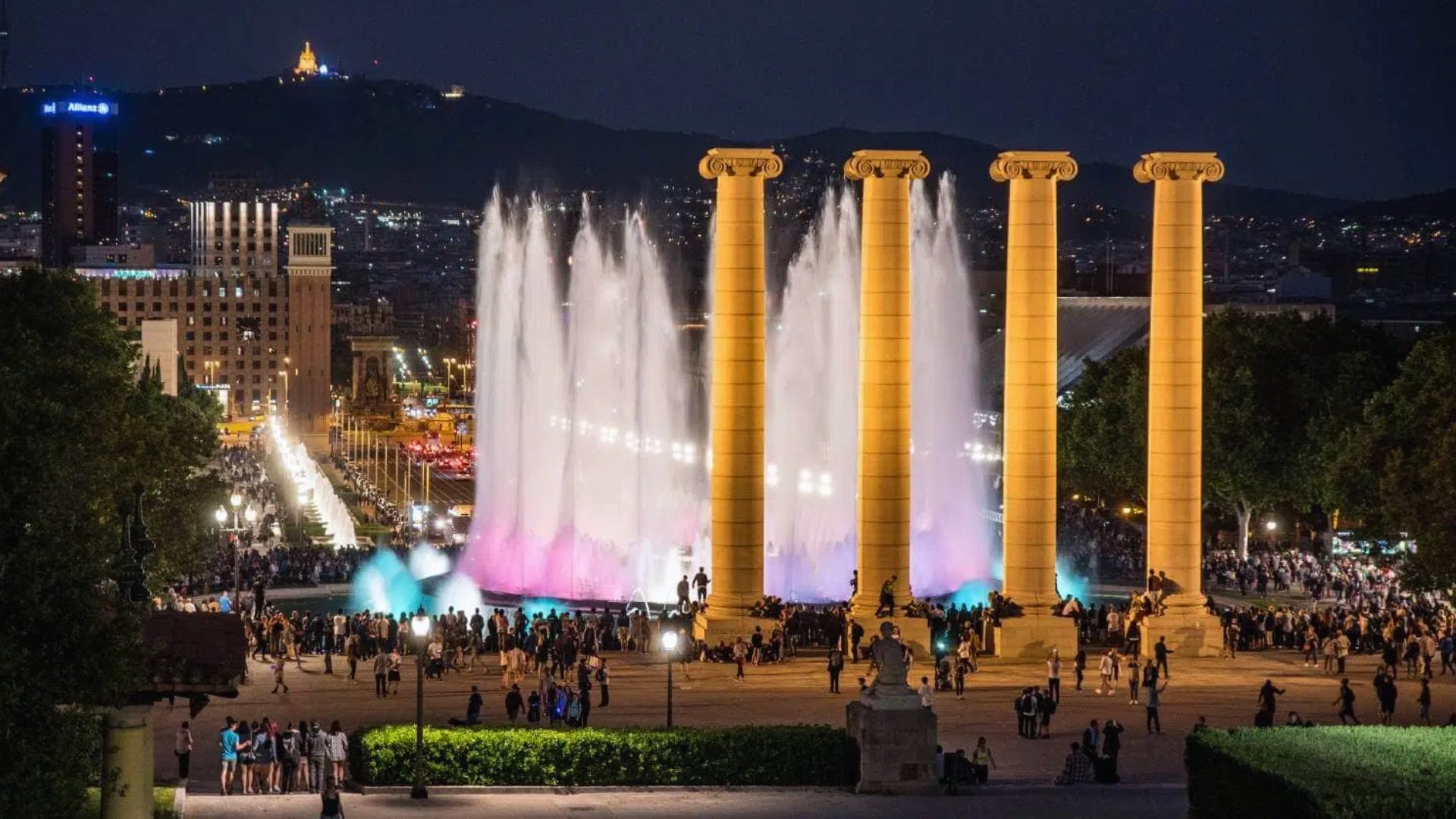 Night scene of illuminated fountains and four tall columns at Montjuïc, with crowds of people and city lights in the background, creating a lively atmosphere.