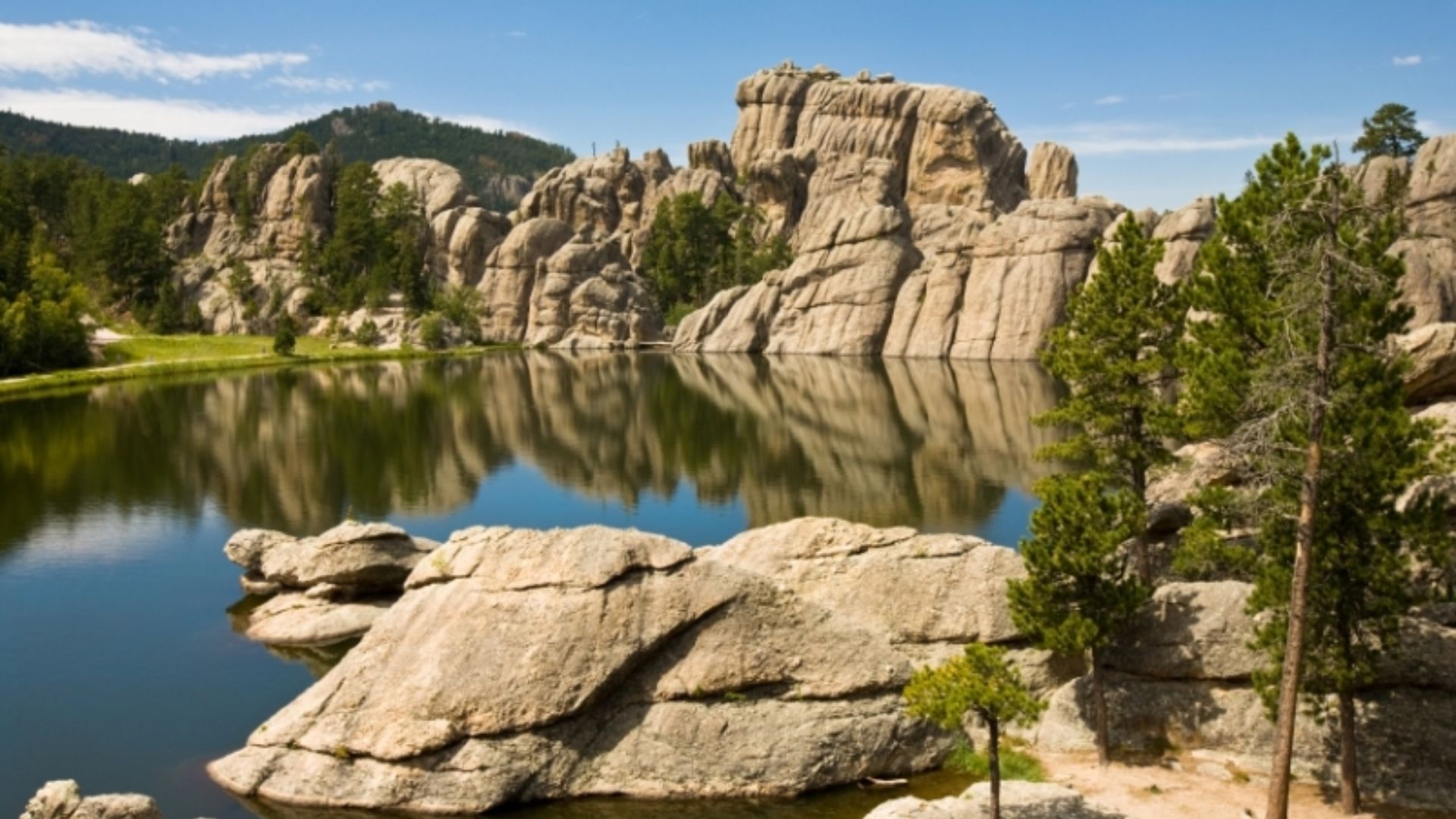 Serene lake scene with large rock formations reflected in the water, surrounded by lush green pine trees under a clear blue sky. Peaceful and scenic.