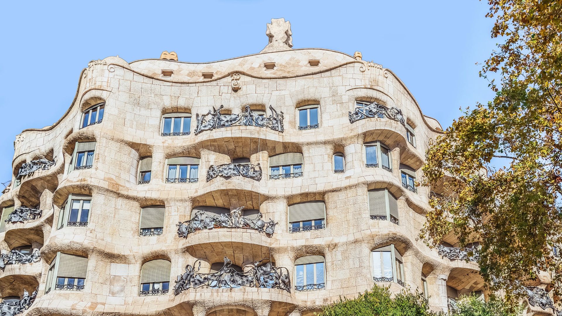 Wavy stone facade of an art nouveau building with ornate metal balconies and irregular windows against a clear blue sky, exuding a whimsical charm.