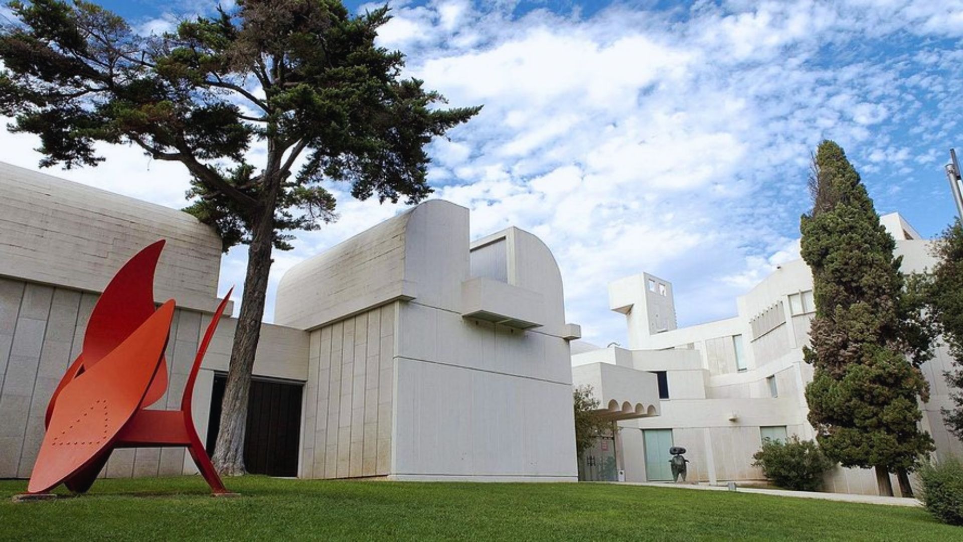 Modern art museum with white, geometric architecture under a blue sky. Features vibrant red abstract sculpture and lush green lawn with trees.