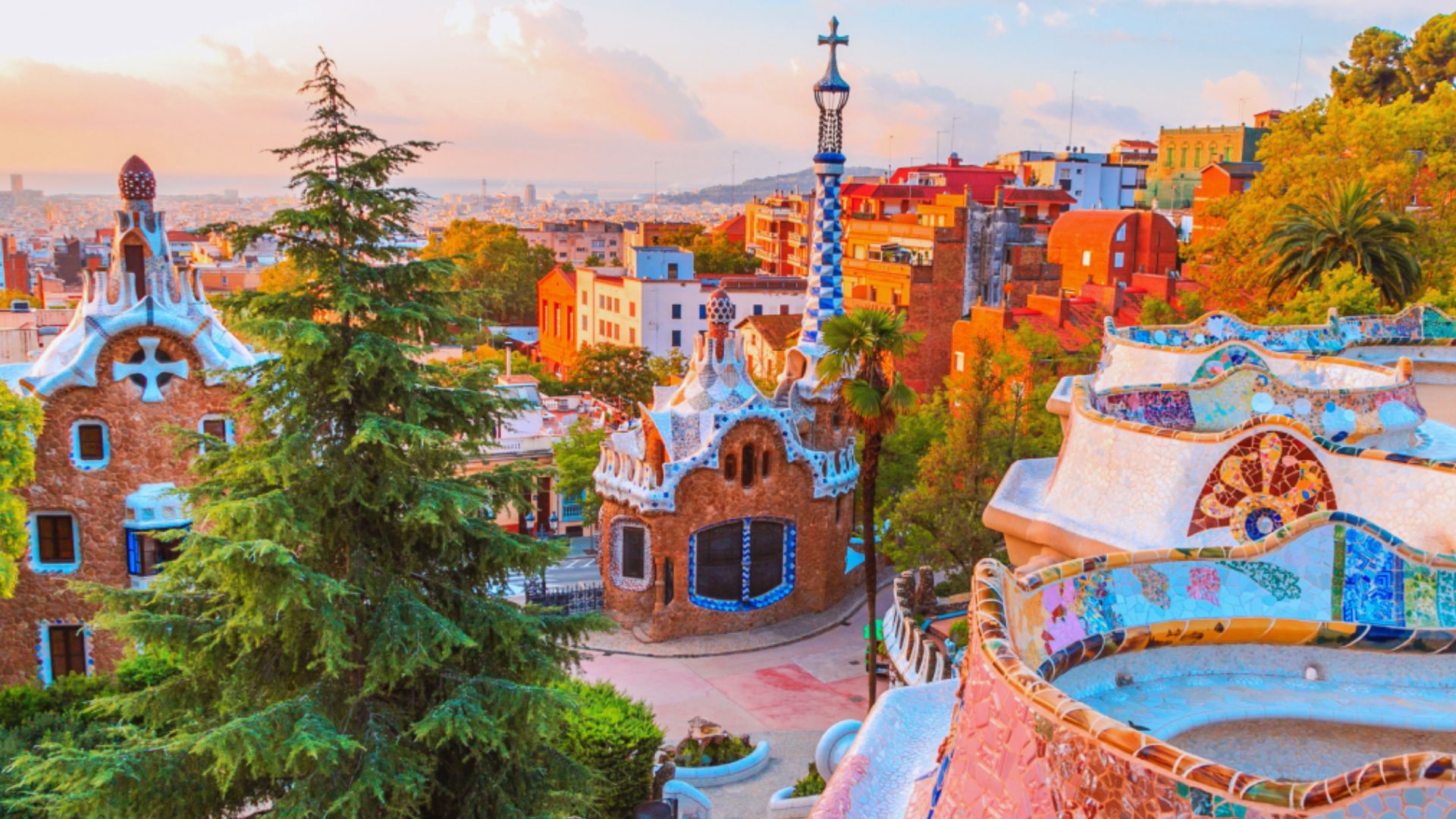 Vibrant view of Park Güell in Barcelona at sunset, showcasing Gaudí's colorful mosaic buildings, lush trees, and a distant cityscape under a pastel sky.