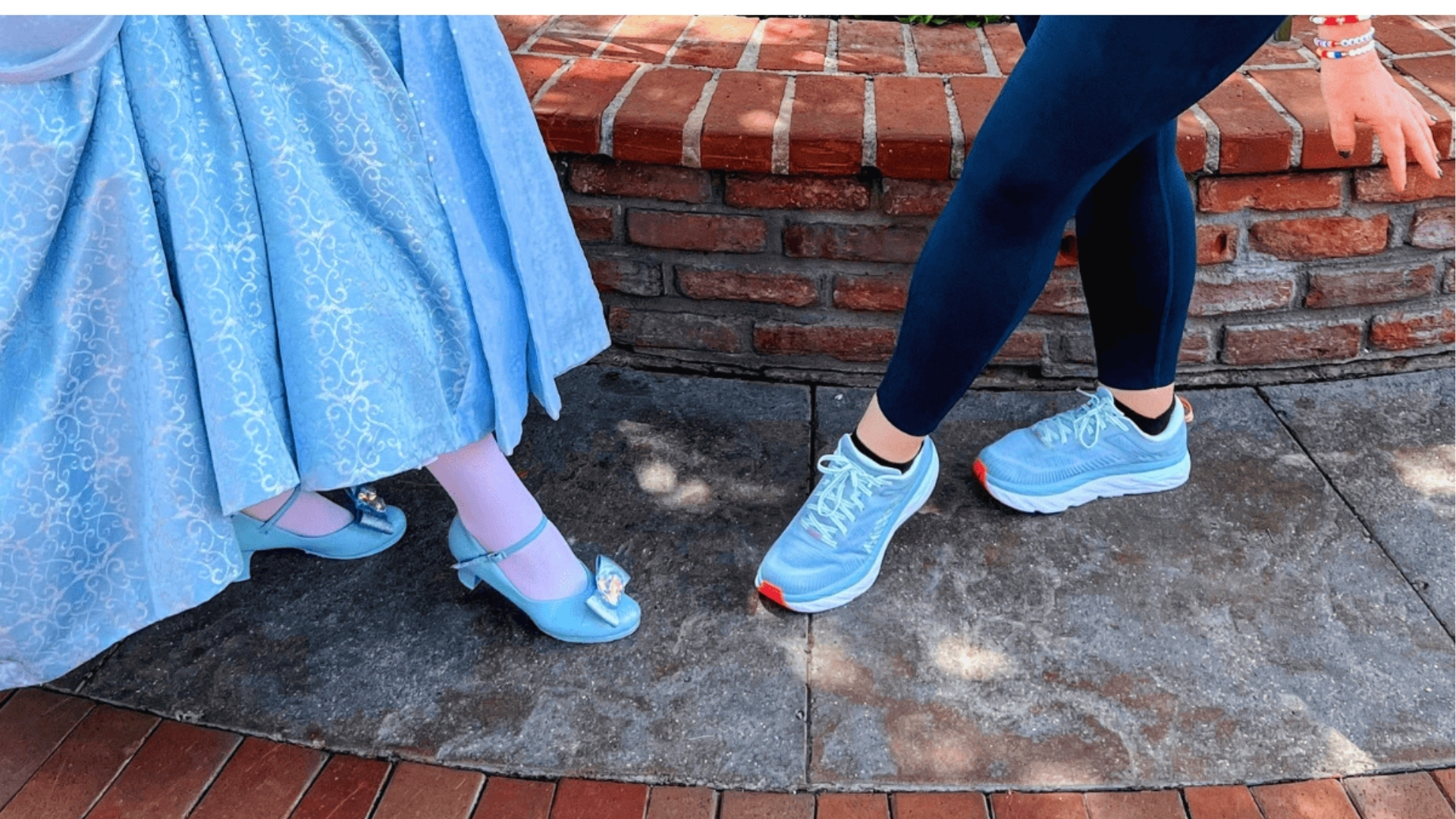 A person in a sparkling blue dress and heels forms a playful pose with someone wearing blue athletic shoes and dark leggings, against a brick wall backdrop.