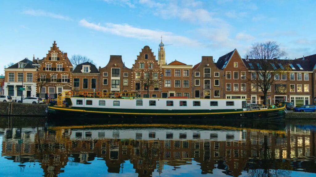 A long canal boat floats on a calm river, reflecting against historic Dutch gabled houses under a blue sky, evoking a serene, picturesque scene.