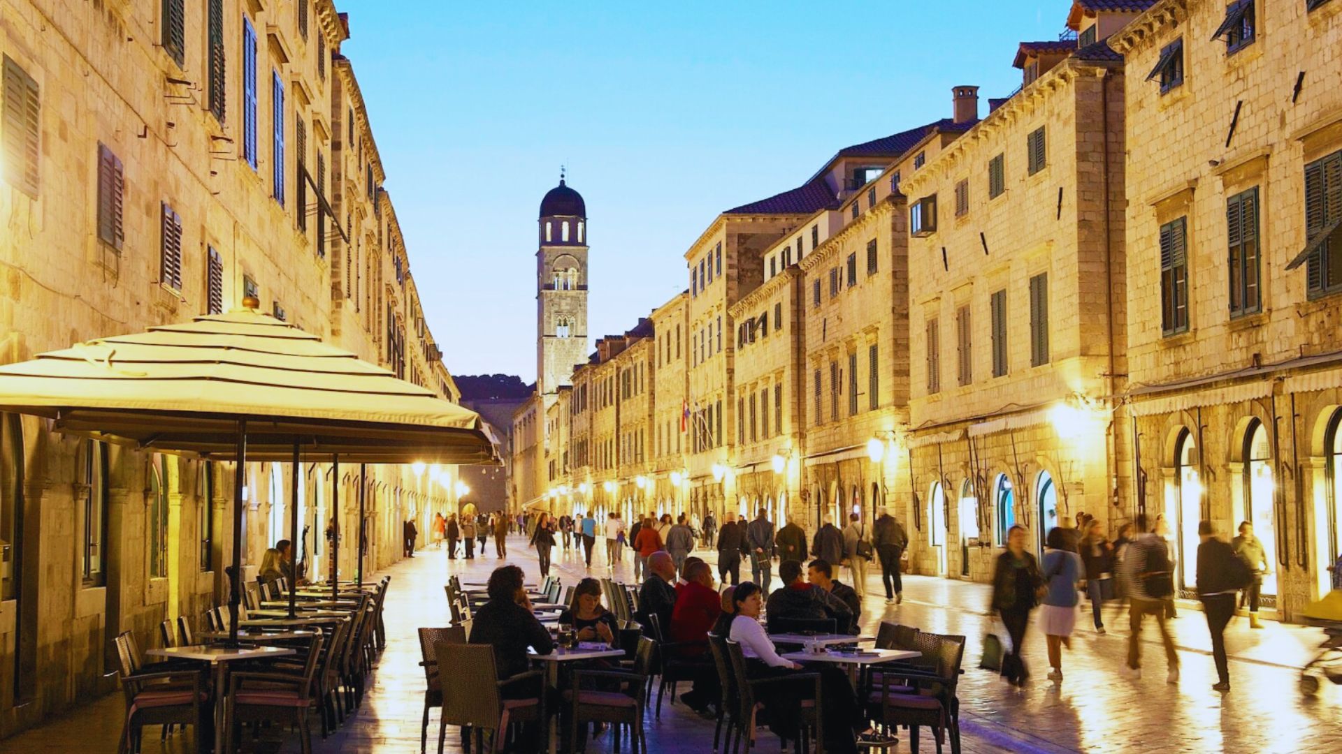 A bustling European street with historic stone buildings and a clock tower at dusk. People walk and dine outdoors, creating a lively and warm atmosphere.