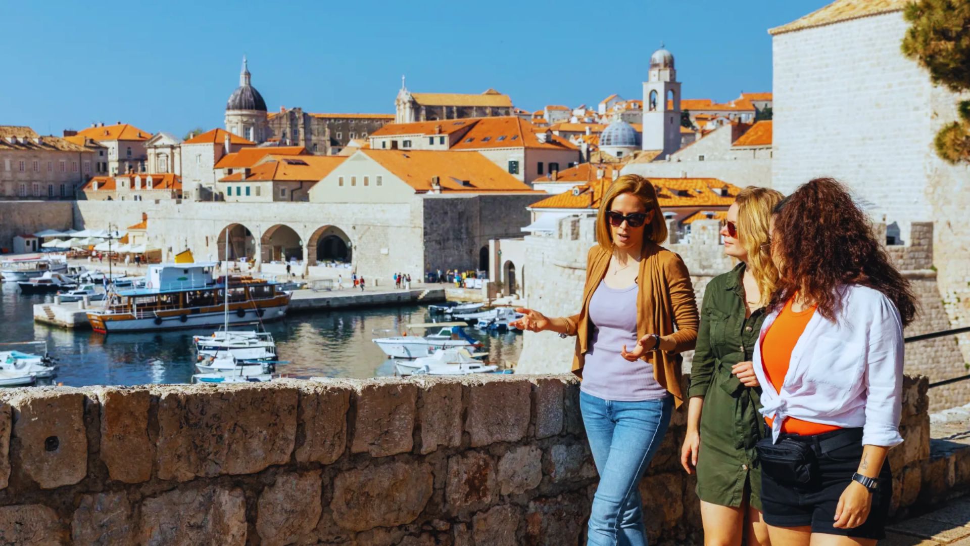 Three women walk along a stone wall overlooking a harbor in Dubrovnik, with historic buildings and orange rooftops in the background under a clear sky.