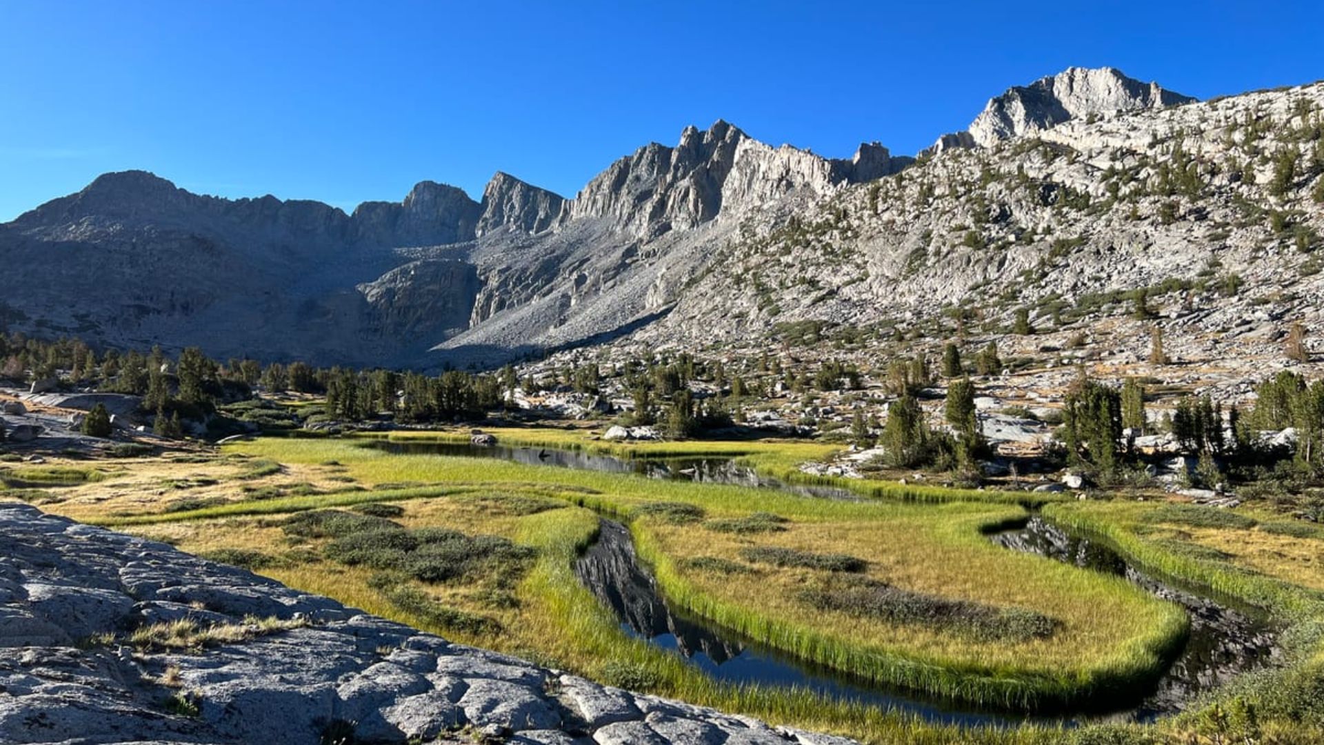 A serene mountain landscape with rugged peaks under a clear blue sky. In the foreground, a lush green meadow surrounds a winding stream, contrasting with the rocky terrain.