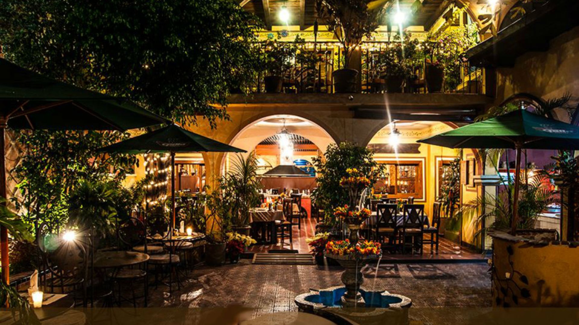 Cozy outdoor restaurant at night featuring warm lighting, lush greenery, and inviting seating. A water fountain sits in the foreground.