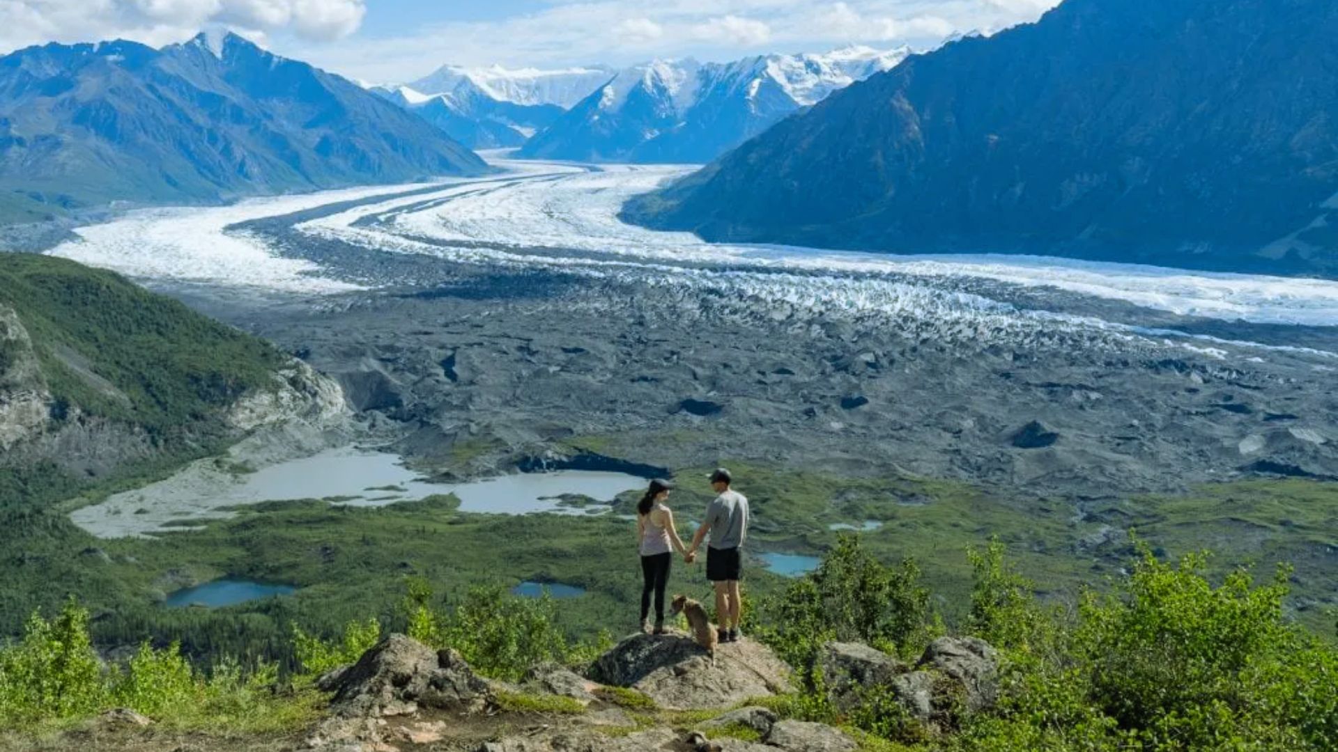 Two individuals stand on a mountain, gazing at a vast glacier below them, surrounded by rugged terrain.