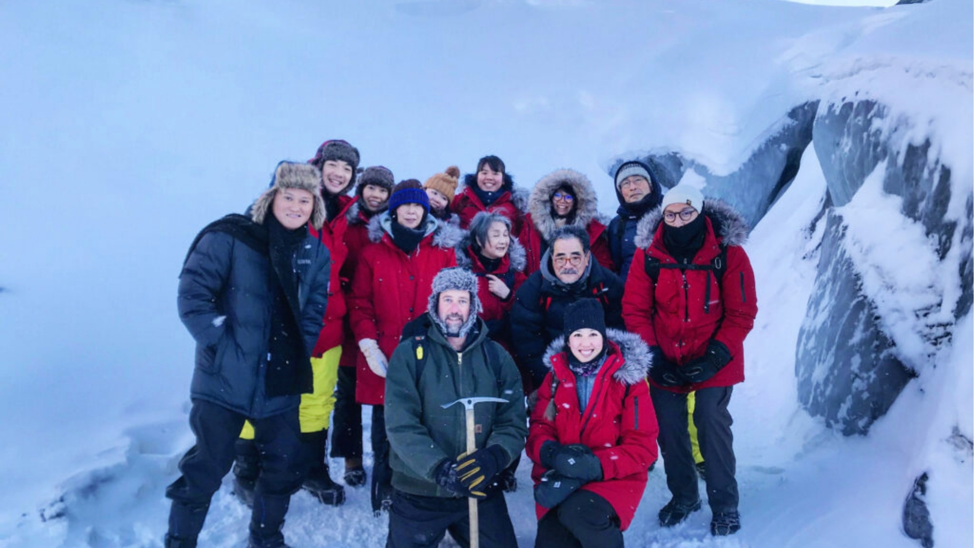 A group of eleven people bundled in winter gear stands on a snowy landscape, smiling. The surrounding area is snow-covered, creating a cold, adventurous atmosphere.