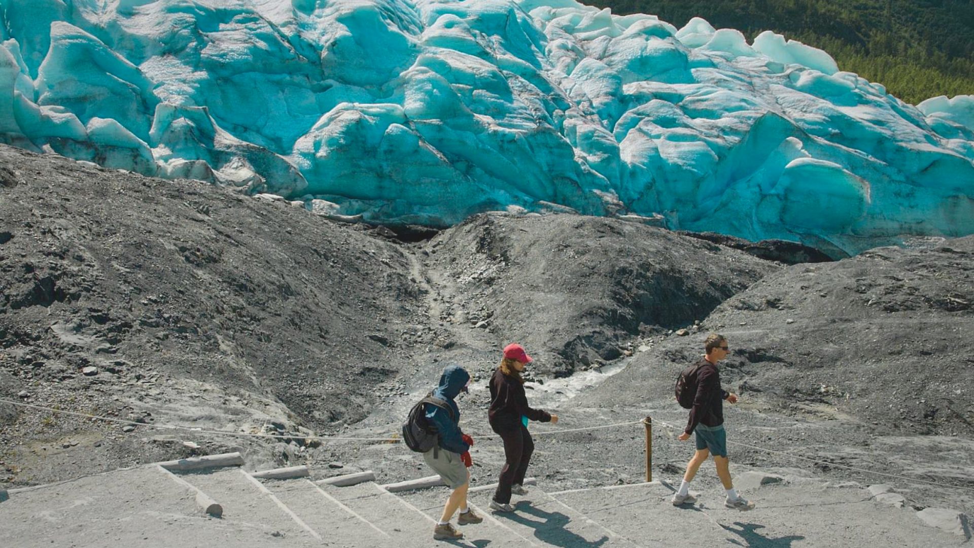 Three people hike down stone steps towards a bright blue glacier, contrasting with the dark rocky terrain. The scene is adventurous and awe-inspiring.