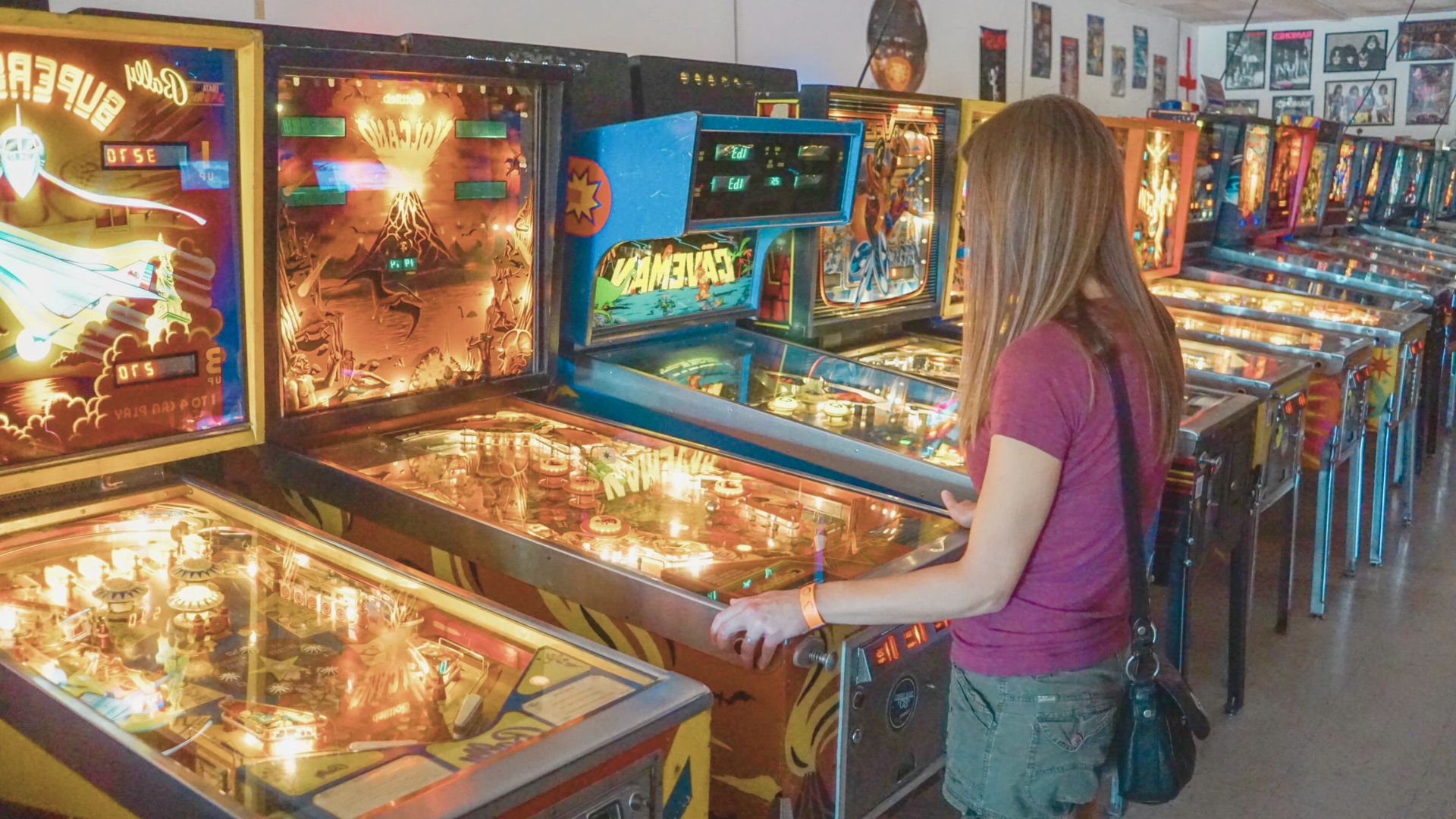 A woman with long hair in a maroon shirt and shorts plays a pinball machine in an arcade filled with colorful vintage machines, evoking nostalgia.