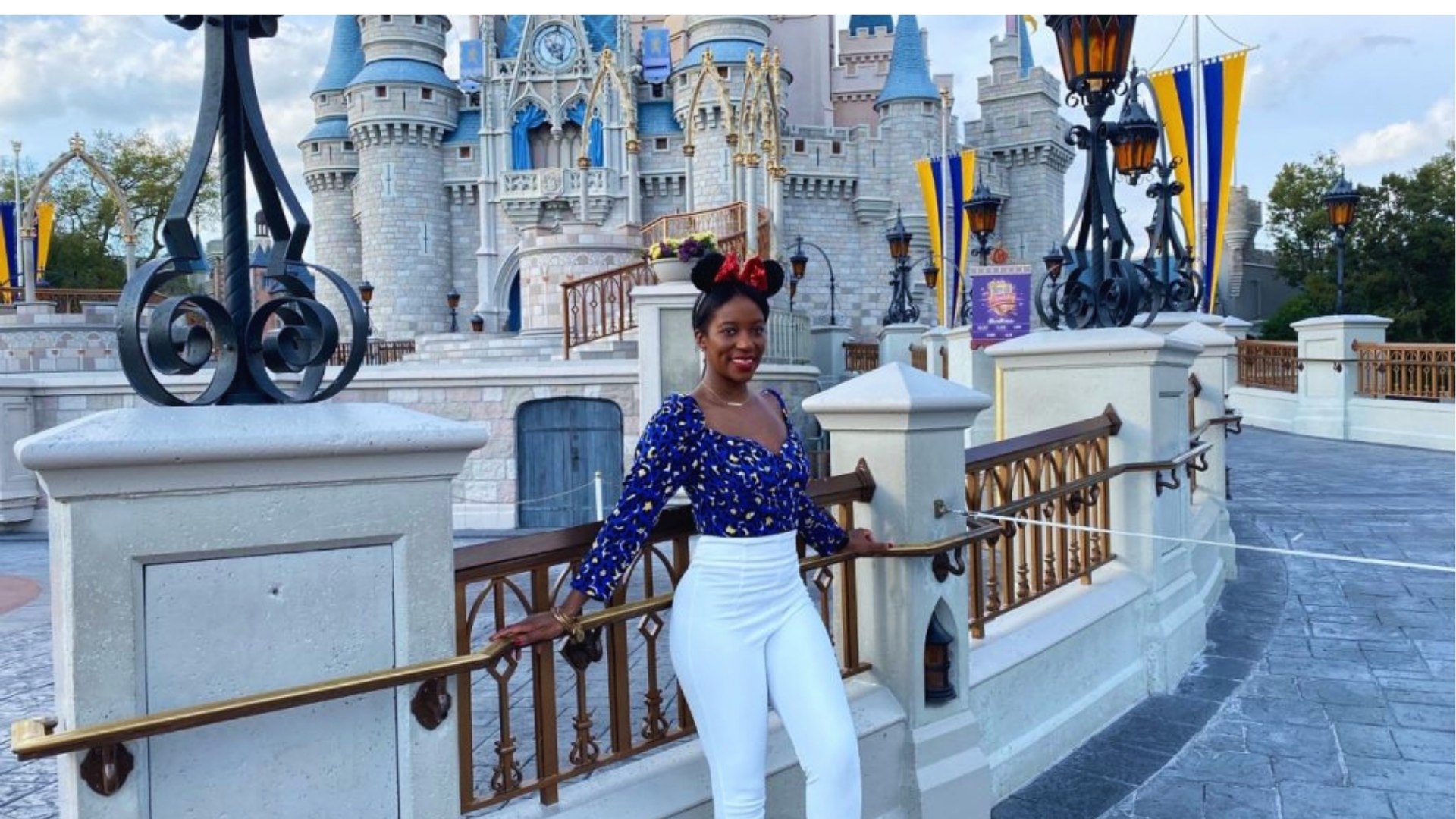 A smiling person with Minnie Mouse ears poses in front of a castle at a theme park. They wear a blue blouse and white pants. The scene is bright and cheerful.
