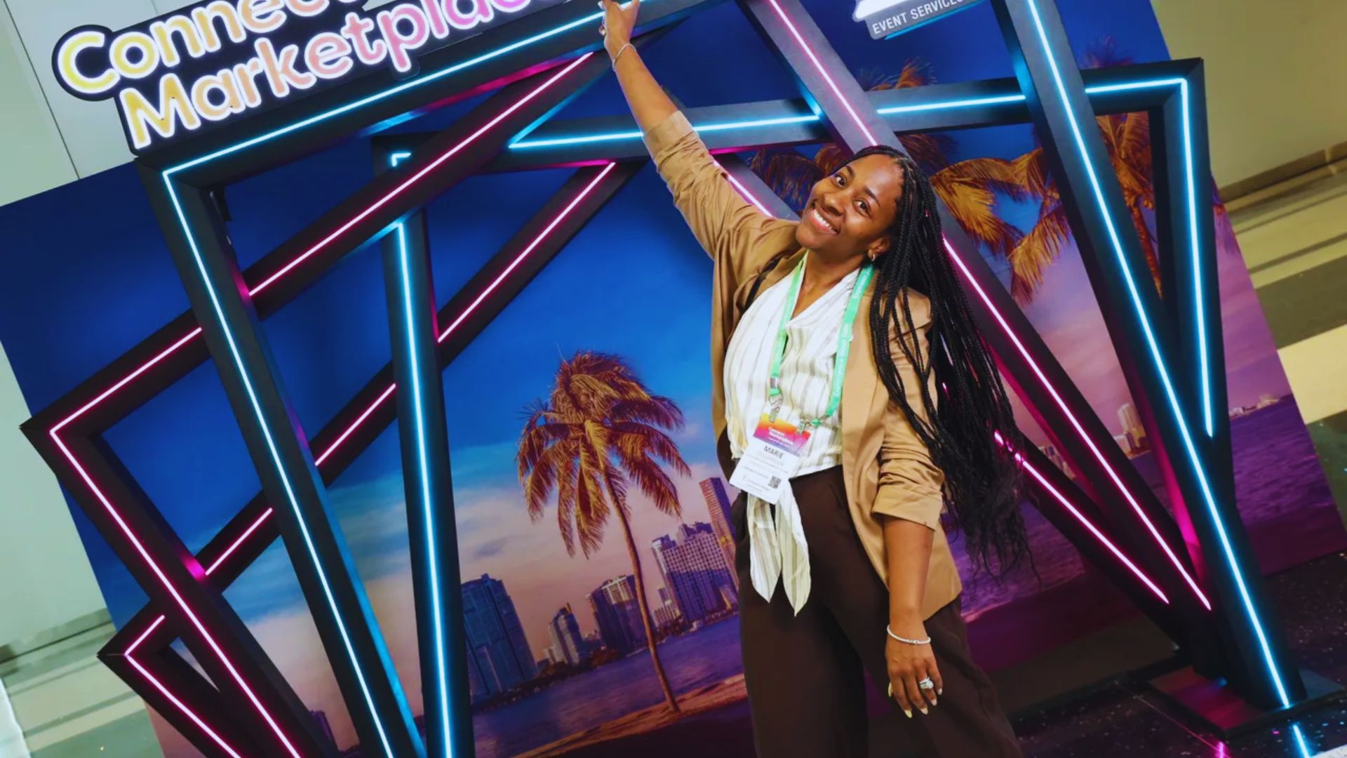 A smiling woman poses energetically in front of a colorful neon frame with palm trees and a city skyline backdrop. The mood is lively and cheerful.