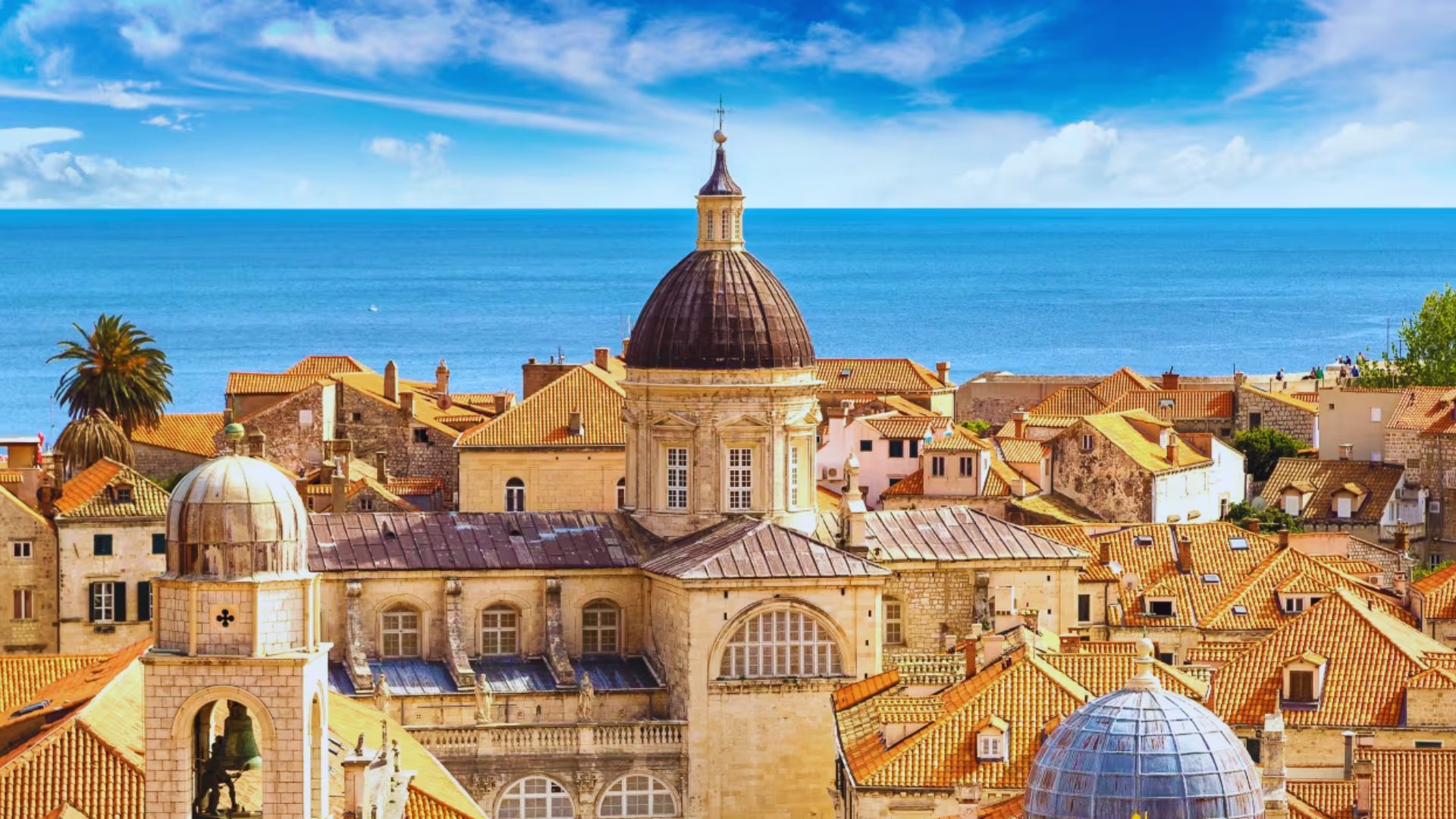 Rooftop view of historic Dubrovnik with terracotta tiles and a prominent domed cathedral. The Adriatic Sea and a vibrant blue sky in the background.