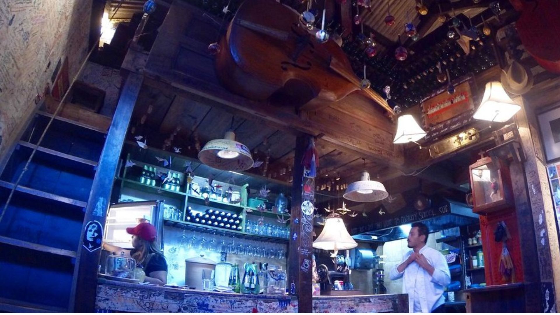 Cozy, eclectic bar with warm lighting, displaying a large cello on the ceiling. A bartender serves drinks, while a man adjusts his shirt nearby.