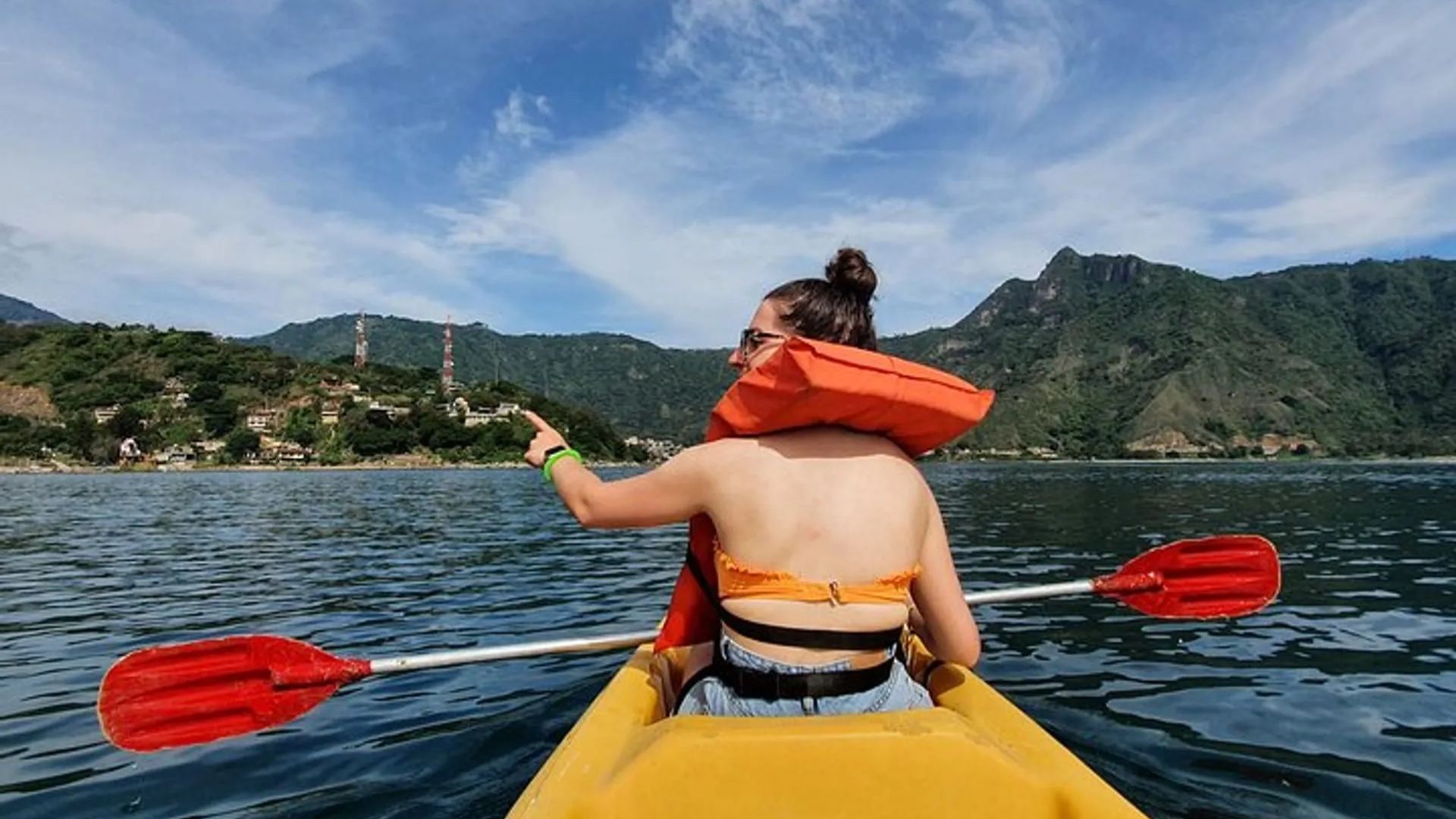 A person in an orange life jacket kayaks on a calm lake with red paddles, surrounded by green hills and a partly cloudy blue sky, pointing left.