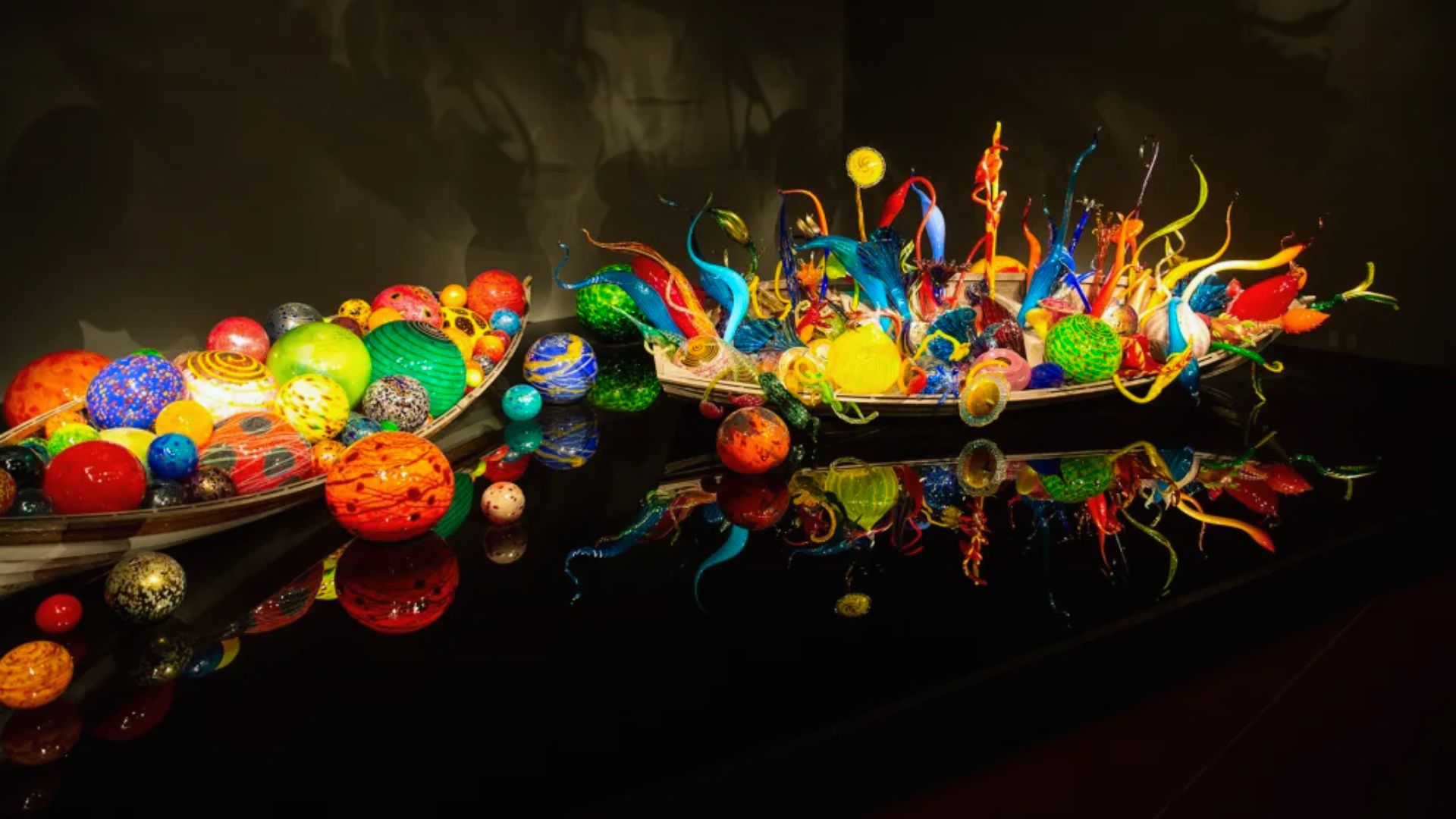 A vivid Chihuly glass sculpture featuring colorful orbs and tendrils reflects on a shiny surface. The art piece is lively and intricate, set against a dark background.