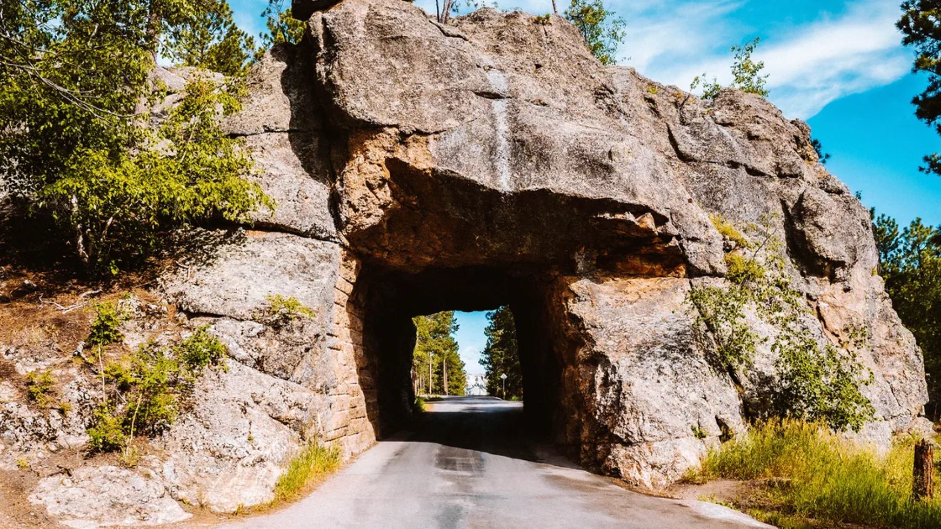 A narrow road passes through a natural rock tunnel surrounded by greenery under a clear blue sky, conveying a sense of adventure and tranquility.