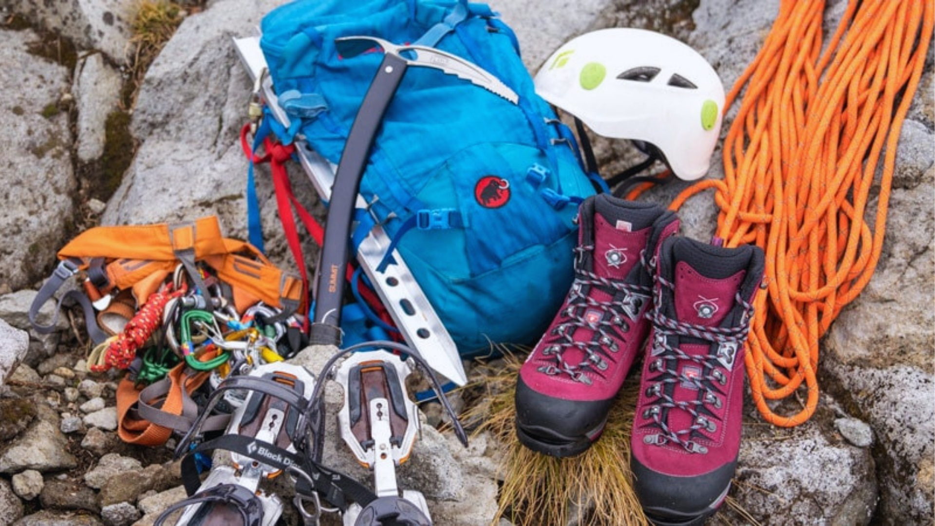A collection of mountaineering gear on rocky ground, including a blue backpack, red boots, white helmet, orange rope, ice axes, and crampons.