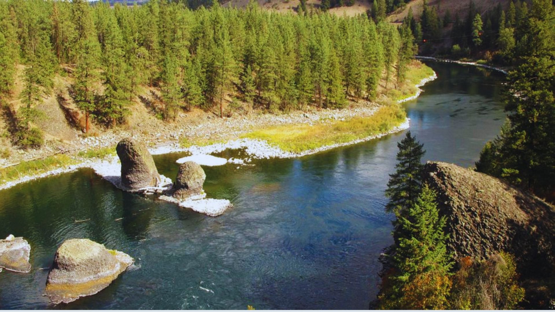A serene river flows through a landscape of lush trees and scattered rocks, creating a tranquil natural scene.
