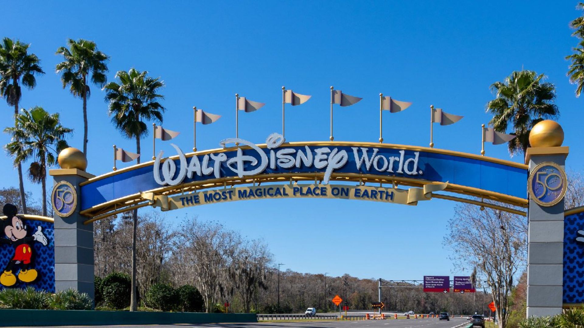 Entrance to Walt Disney World featuring a blue arch with gold accents and flags, flanked by palm trees. Mickey Mouse is depicted on the left pillar.
