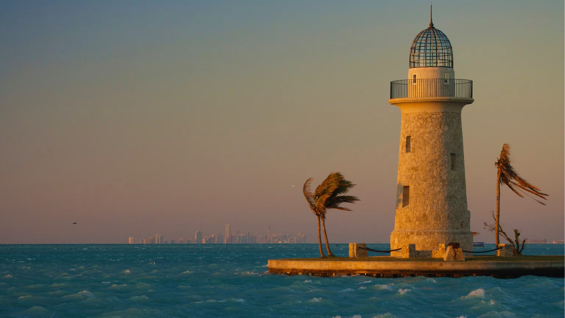 Lighthouse at sunset on a pier with swaying palm trees, overlooking a calm teal ocean. Distant city skyline under a pink-orange sky creates a tranquil scene.