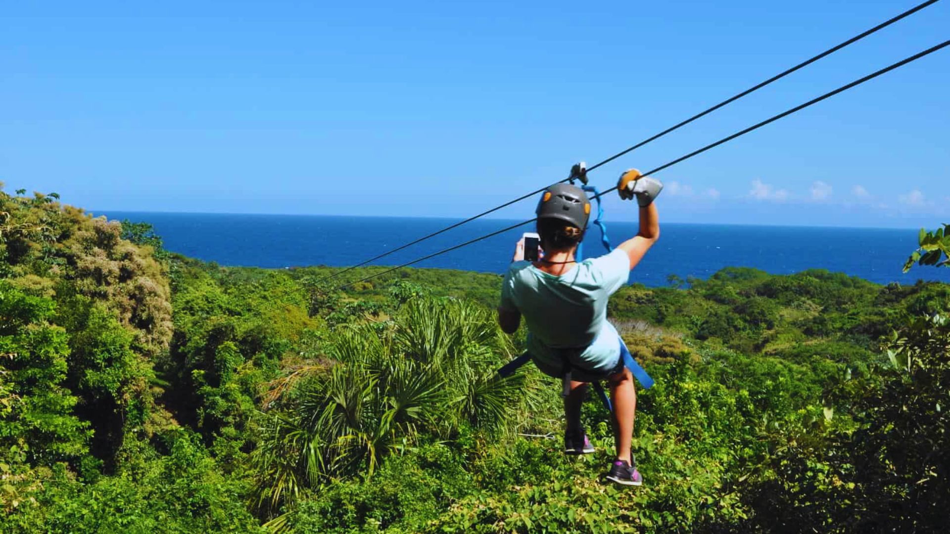 A person zip-lining over lush green forests with a view of the distant blue ocean under a clear sky, conveying a sense of adventure and excitement.