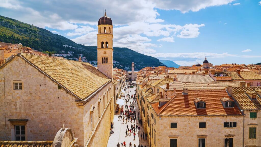 A scenic view of Dubrovnik's Old Town with a tall bell tower, medieval stone buildings, and a busy cobblestone street under a clear blue sky.