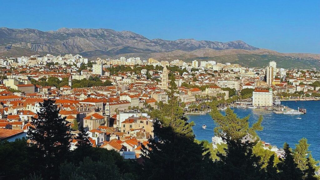 Split vs Dubrovnik: Which Croatian City to Visit? Aerial view of a coastal city with red-roofed buildings, a prominent bell tower, and a bustling harbor. Mountains in the background under a clear blue sky.