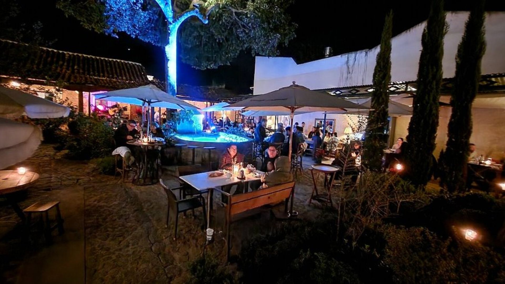 Nighttime outdoor restaurant with warm candlelight on tables, people dining under umbrellas. A large tree glows blue, creating a cozy atmosphere.