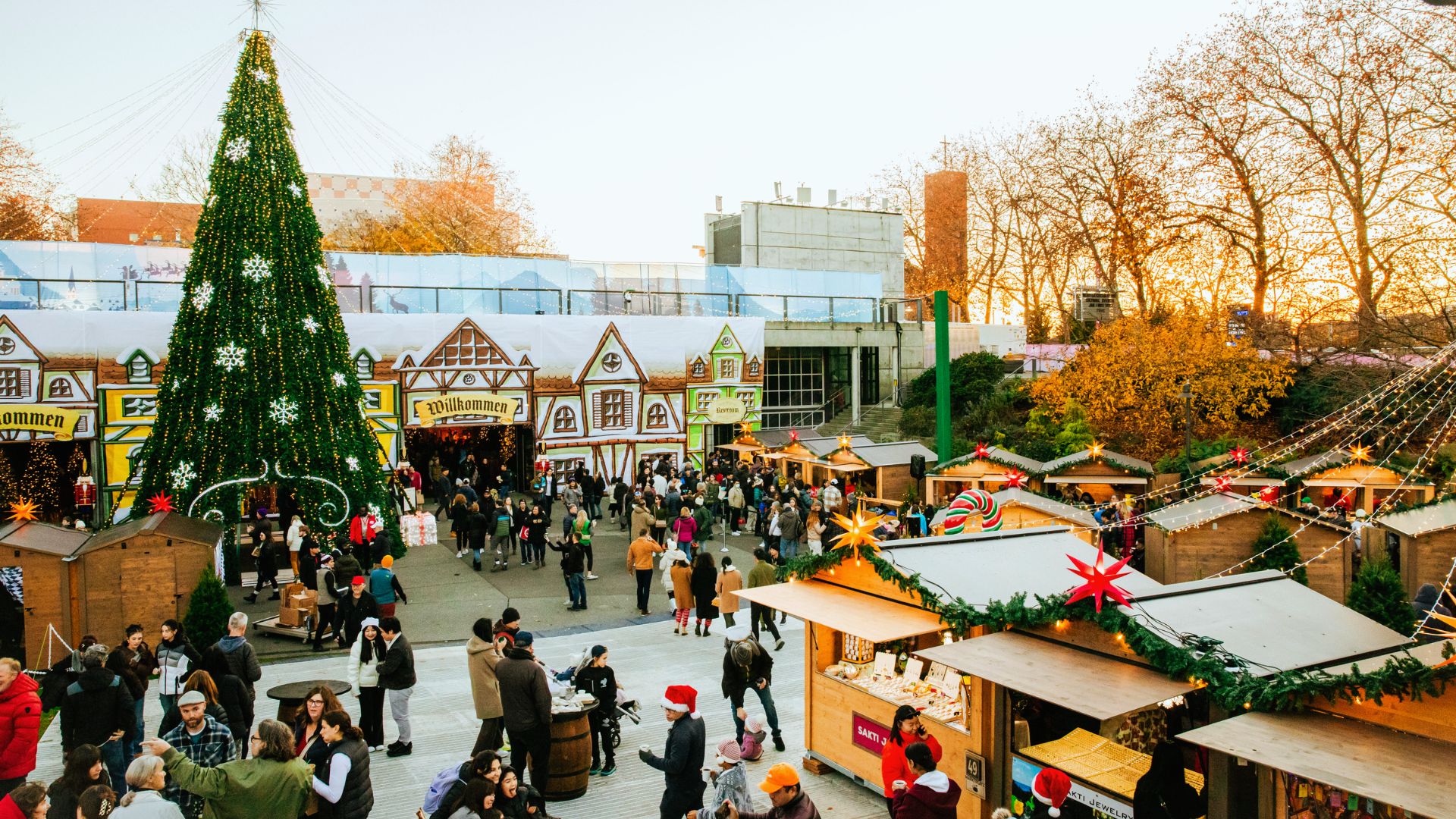 Festive Christmas market with people browsing stalls under colorful lights. A large decorated tree stands on the left, surrounded by lively, cheerful visitors.