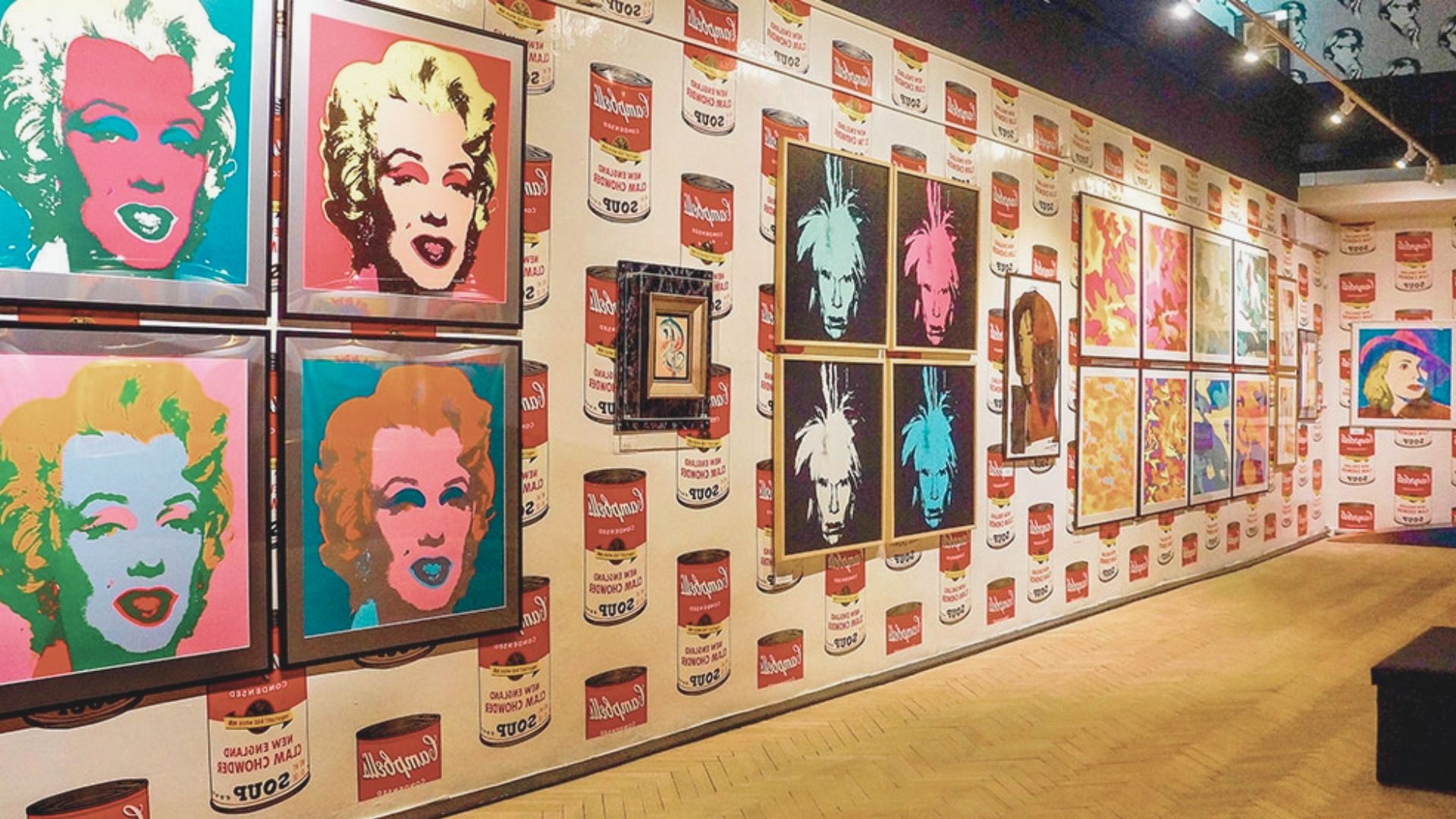 Art gallery with pop art on a wall covered in Campbell's soup can patterns. Features colorful portraits, evoking a vibrant and playful tone.