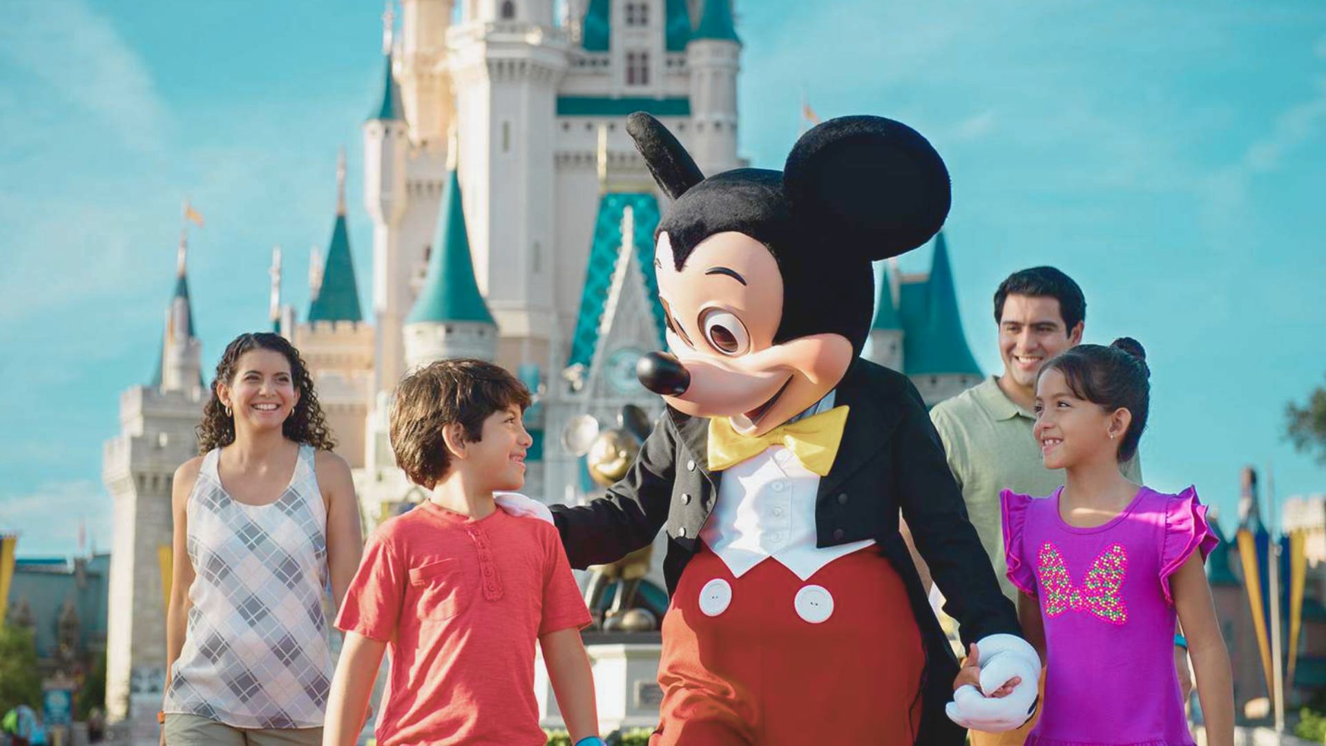 A family smiles joyfully with Mickey Mouse in front of a castle at a theme park. The mood is cheerful and magical under a bright blue sky.