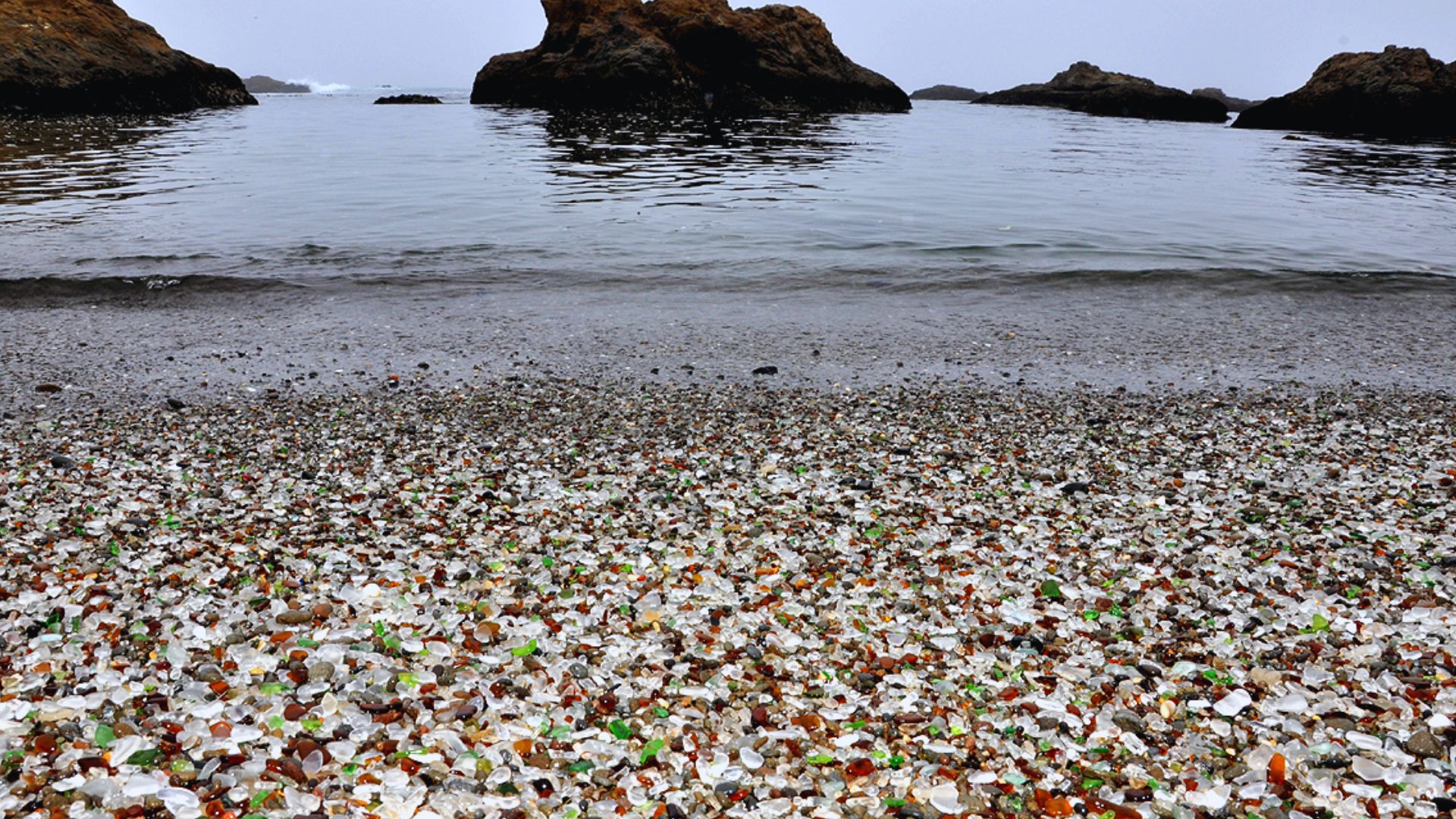 A beach scattered with colorful glass pieces, reflecting sunlight against the sand and waves.