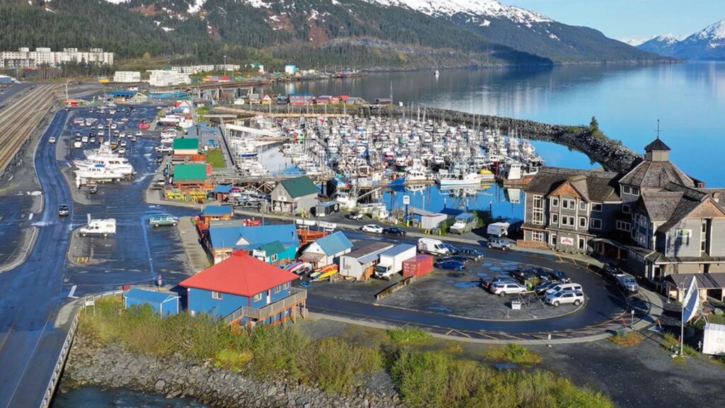 Coastal marina scene with colorful buildings and numerous docked boats. Snow-capped mountains and calm water in the background create a serene atmosphere.