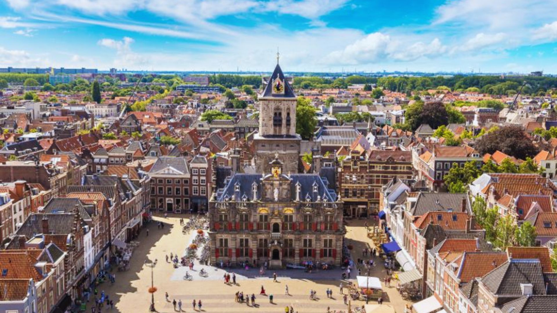 Aerial view of a European city square with a historic central building featuring ornate architecture, surrounded by red-roofed houses under a bright blue sky.
