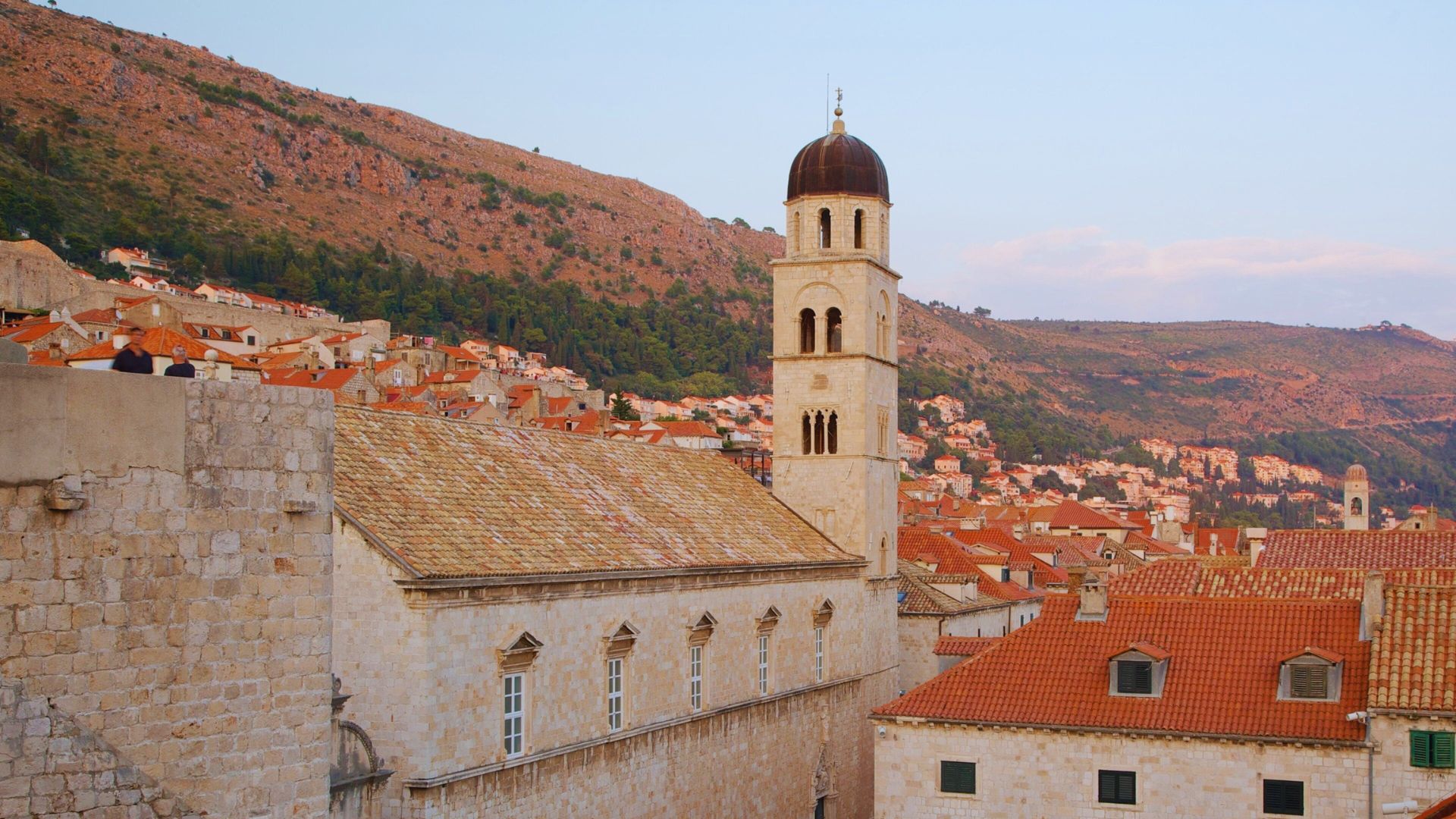 Old town with a prominent bell tower and terracotta rooftops at sunset, nestled against a hillside. Warm, historic atmosphere with serene tones.