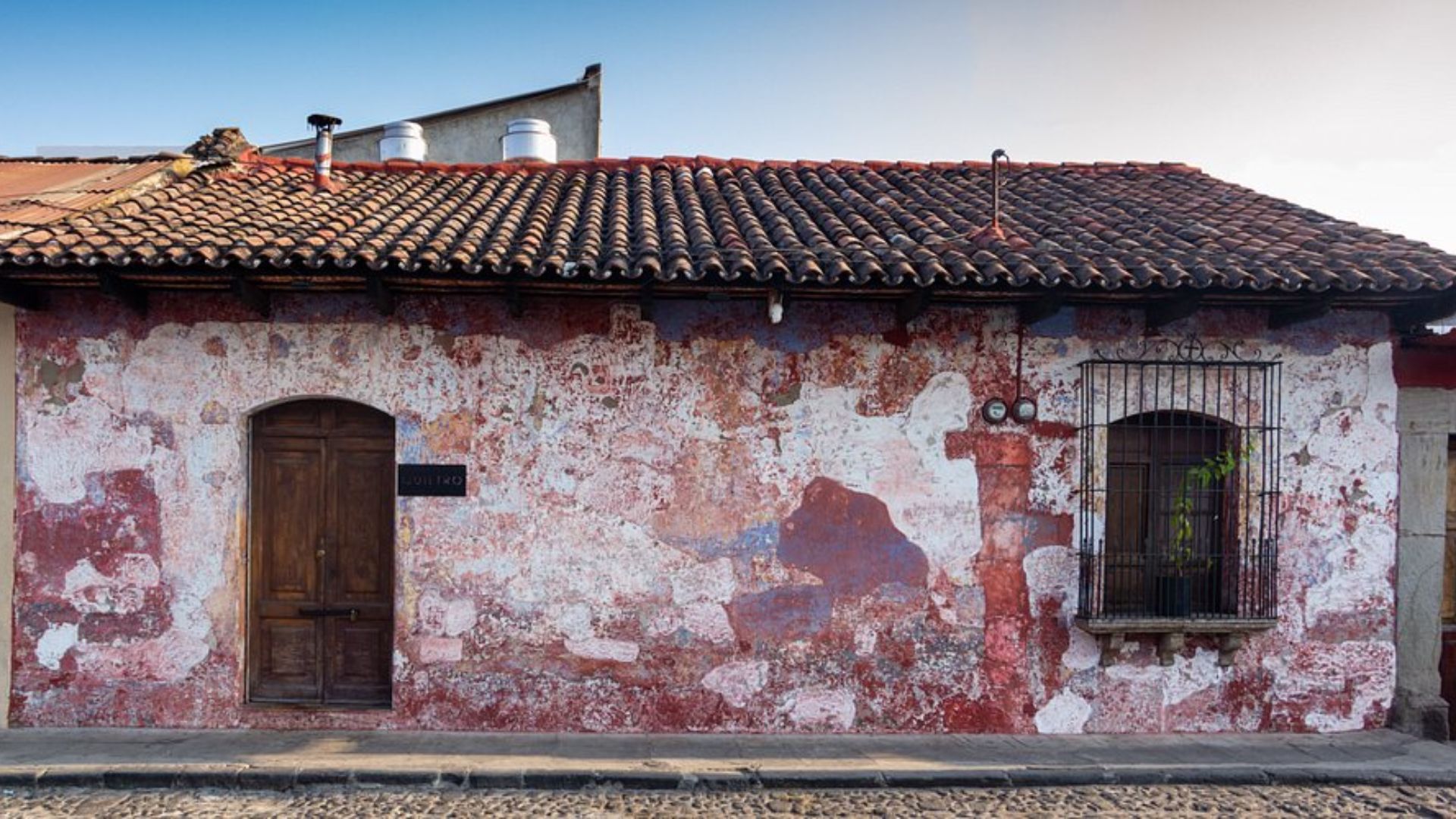 An old, weathered building with peeling red and white paint, a wooden door, and a barred window. The mood conveys rustic charm and historical character.