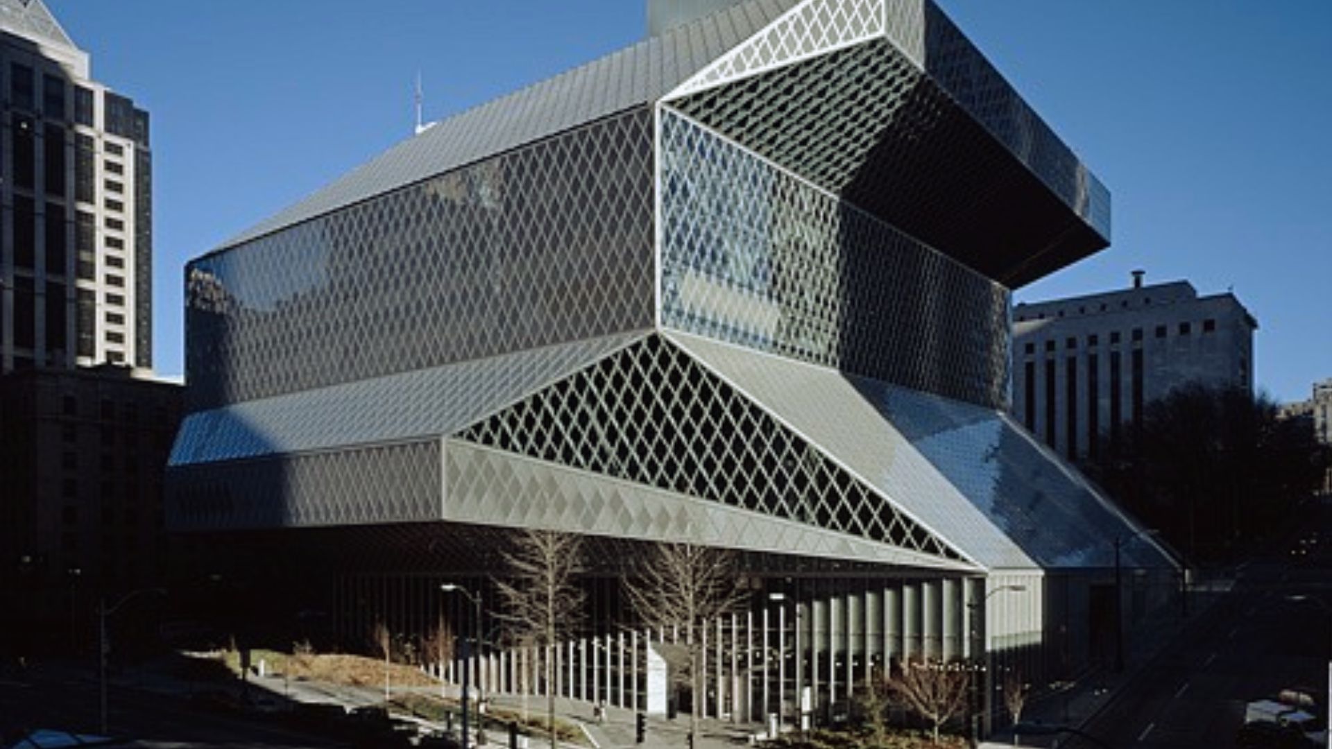 Futuristic library with a geometric design of overlapping metallic and glass panels, surrounded by urban buildings under a clear blue sky.