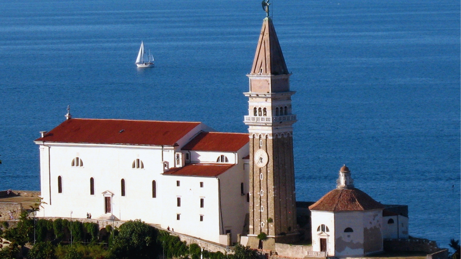 A church with a tower sits atop a hill, overlooking the ocean and surrounded by lush greenery.