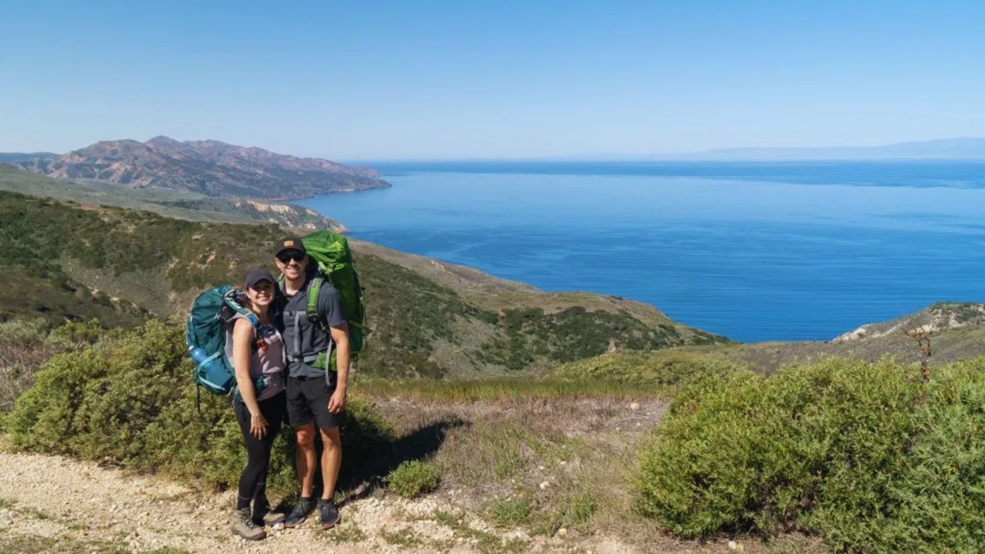 Two hikers with backpacks stand on a trail, gazing at the ocean view below them.