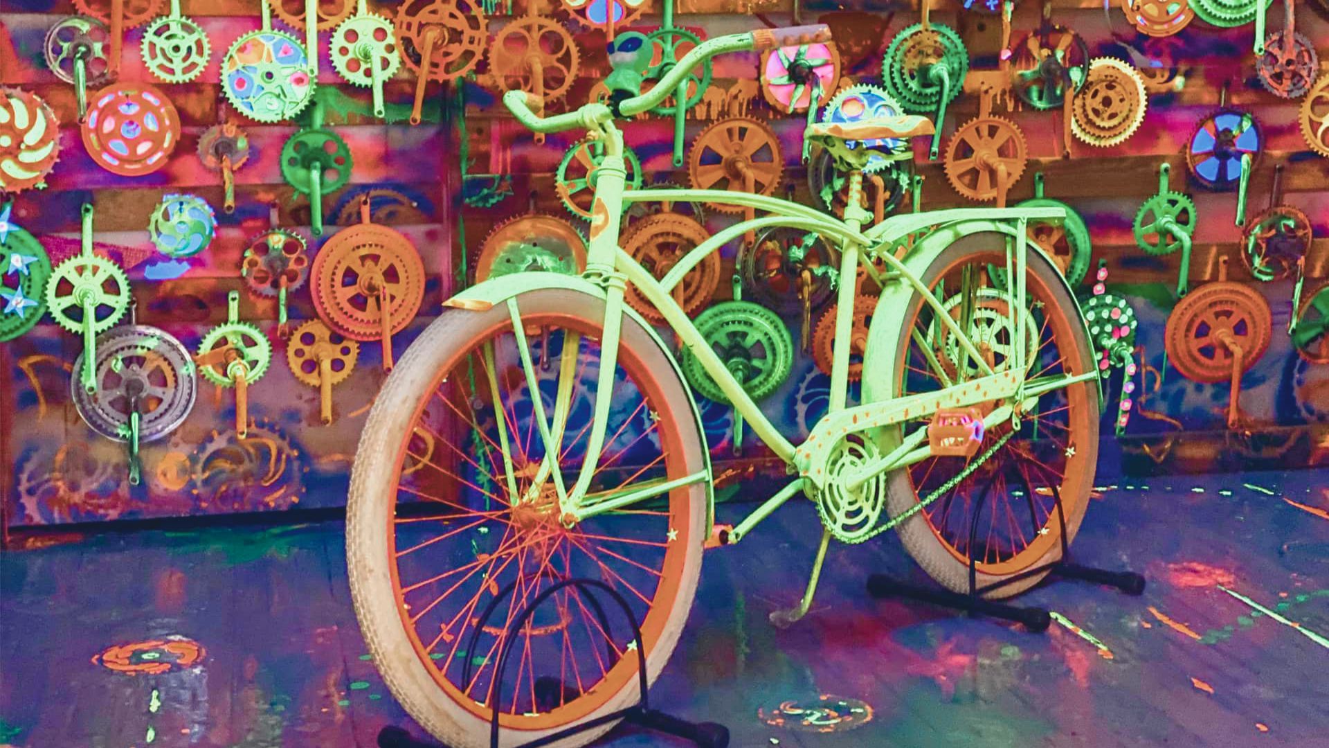 A bright green bicycle stands in front of a colorful wall adorned with variously-sized, vibrant bicycle gears, creating a whimsical, artistic atmosphere.