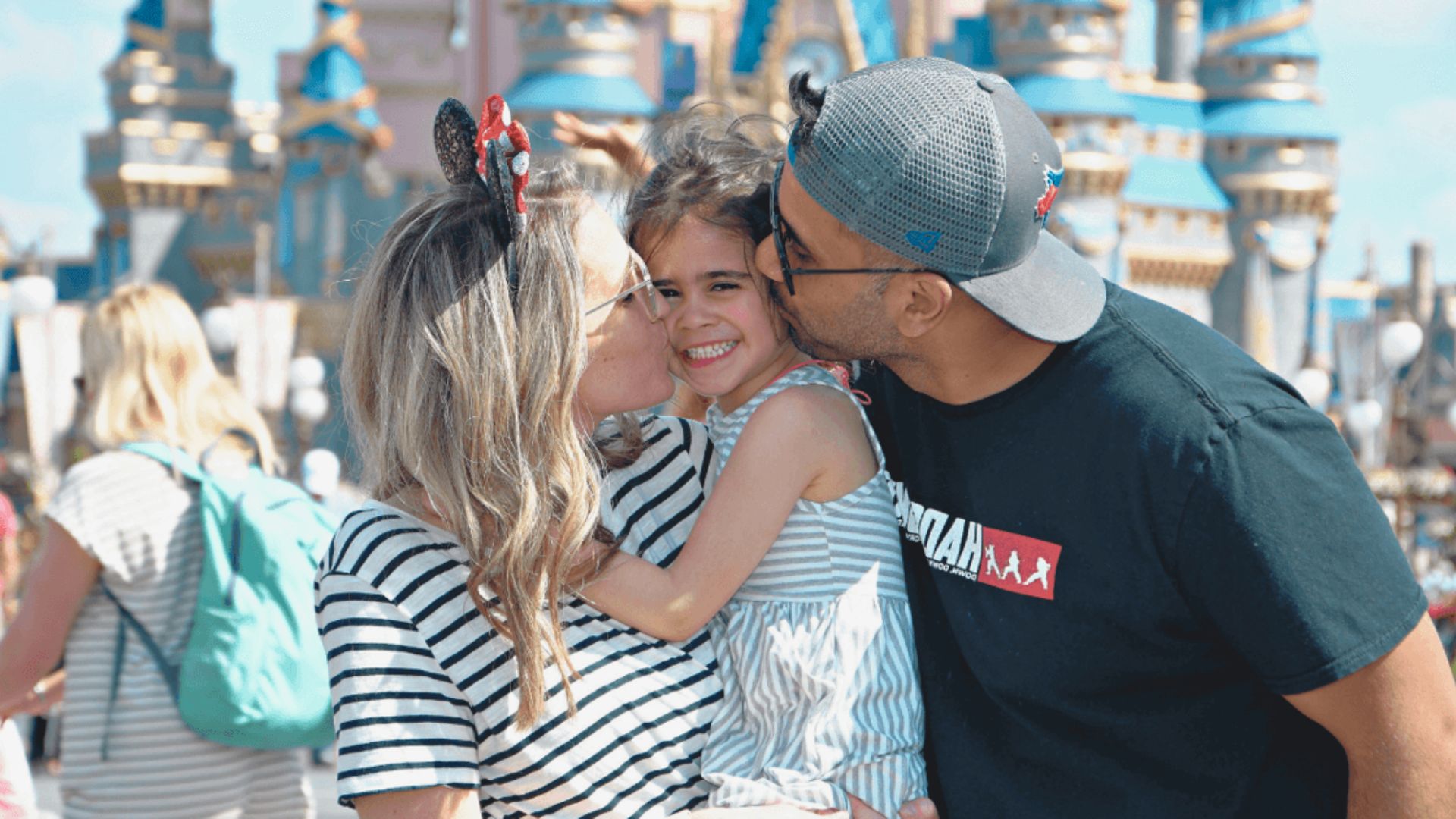 A joyful family at a theme park; parents kiss their smiling daughter, all wearing casual clothes. Background features a castle with blue spires.