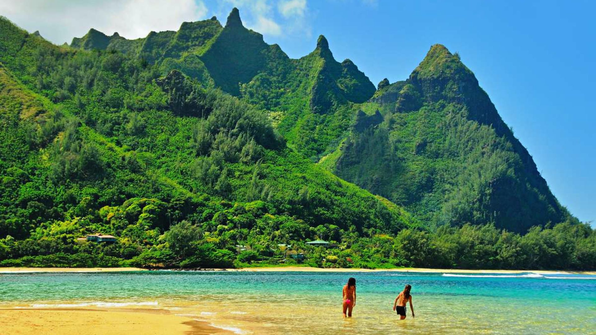 Two people stand in shallow water, with a majestic mountain rising in the background under a clear blue sky.