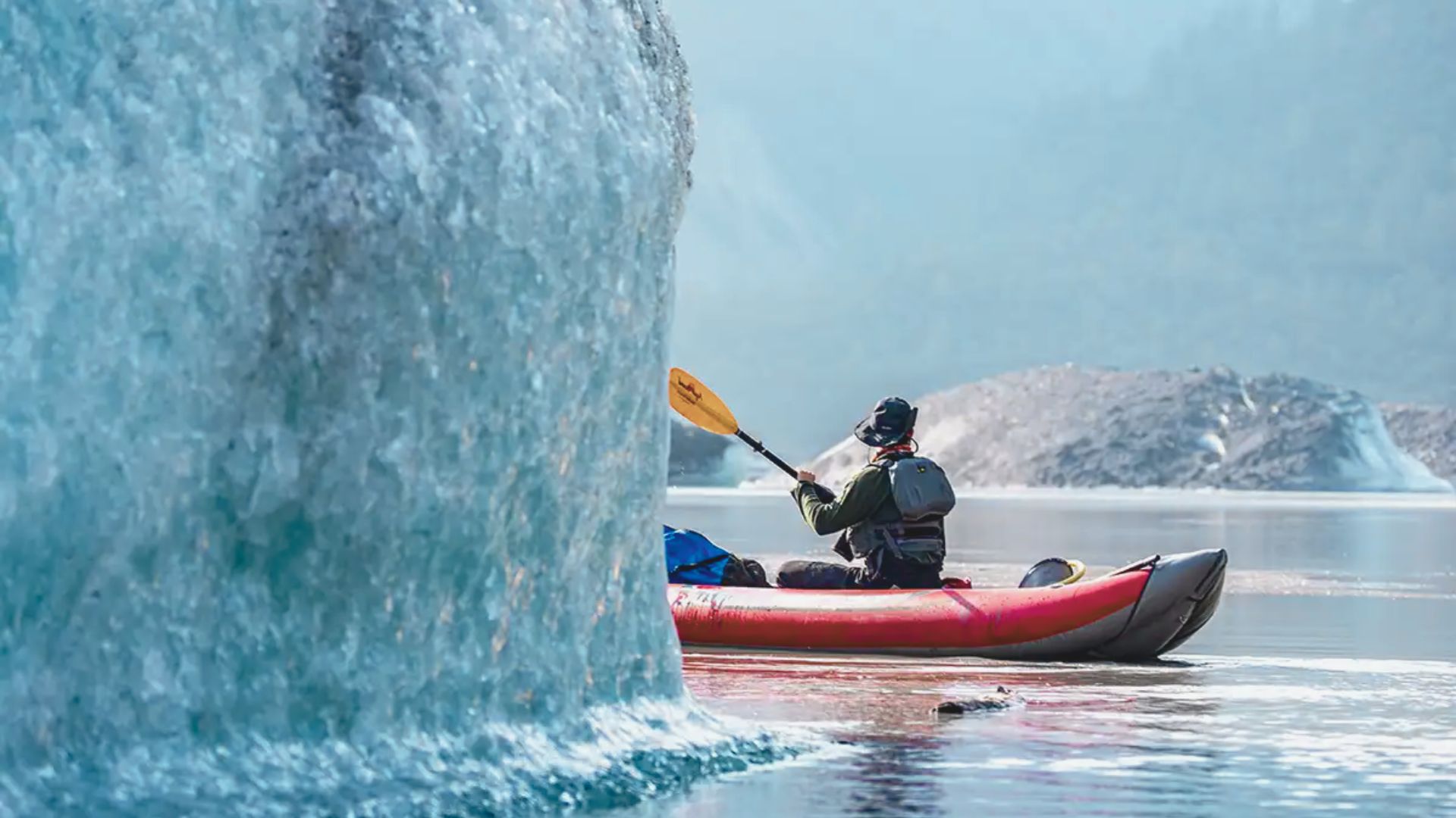 A person in a kayak paddles near a massive ice formation on a calm, clear lake. The scene is serene, with distant, misty mountains visible.