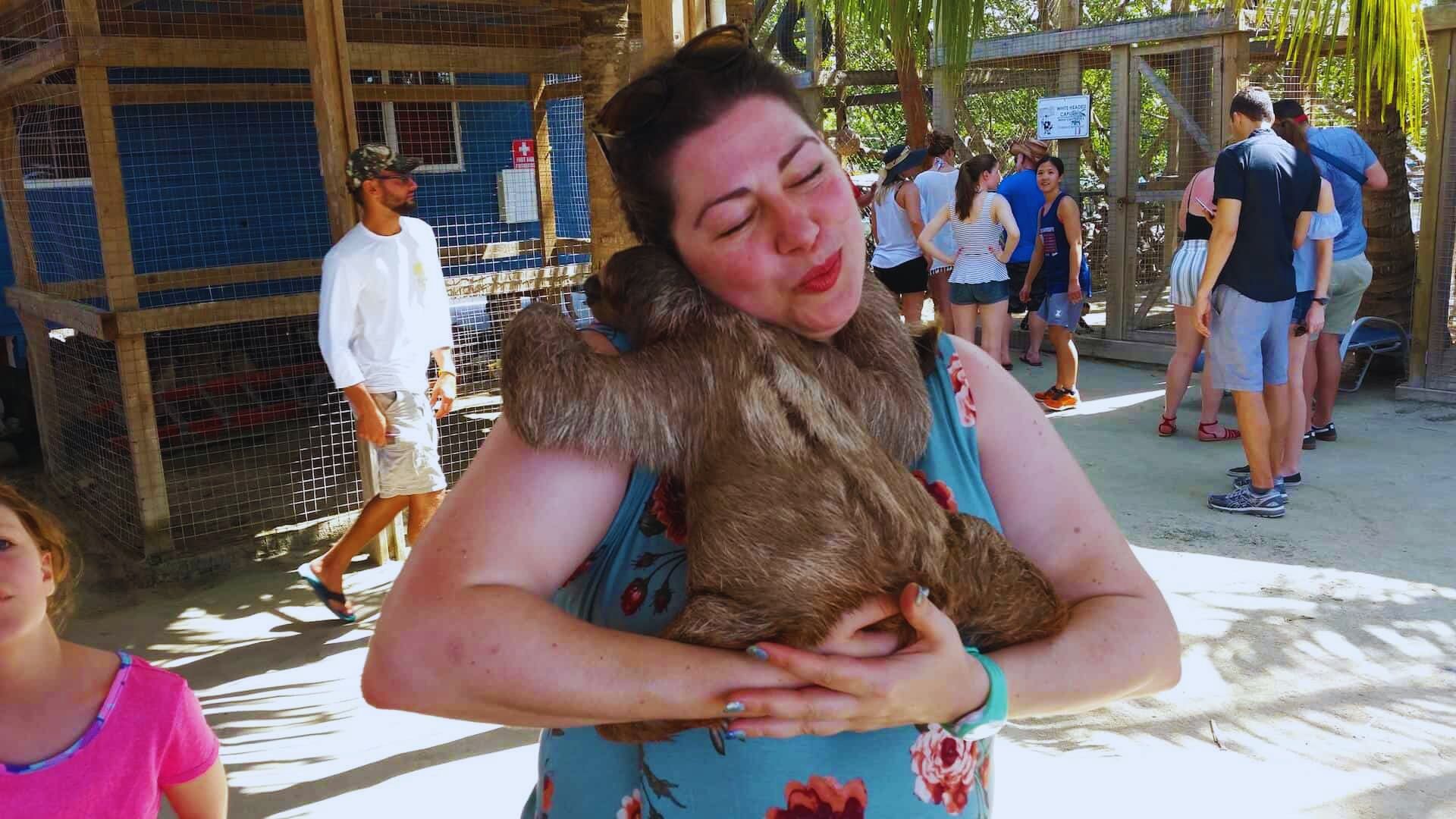 A smiling woman in a floral dress hugs a sloth affectionately at an outdoor animal enclosure. People and trees are blurred in the sunny background.